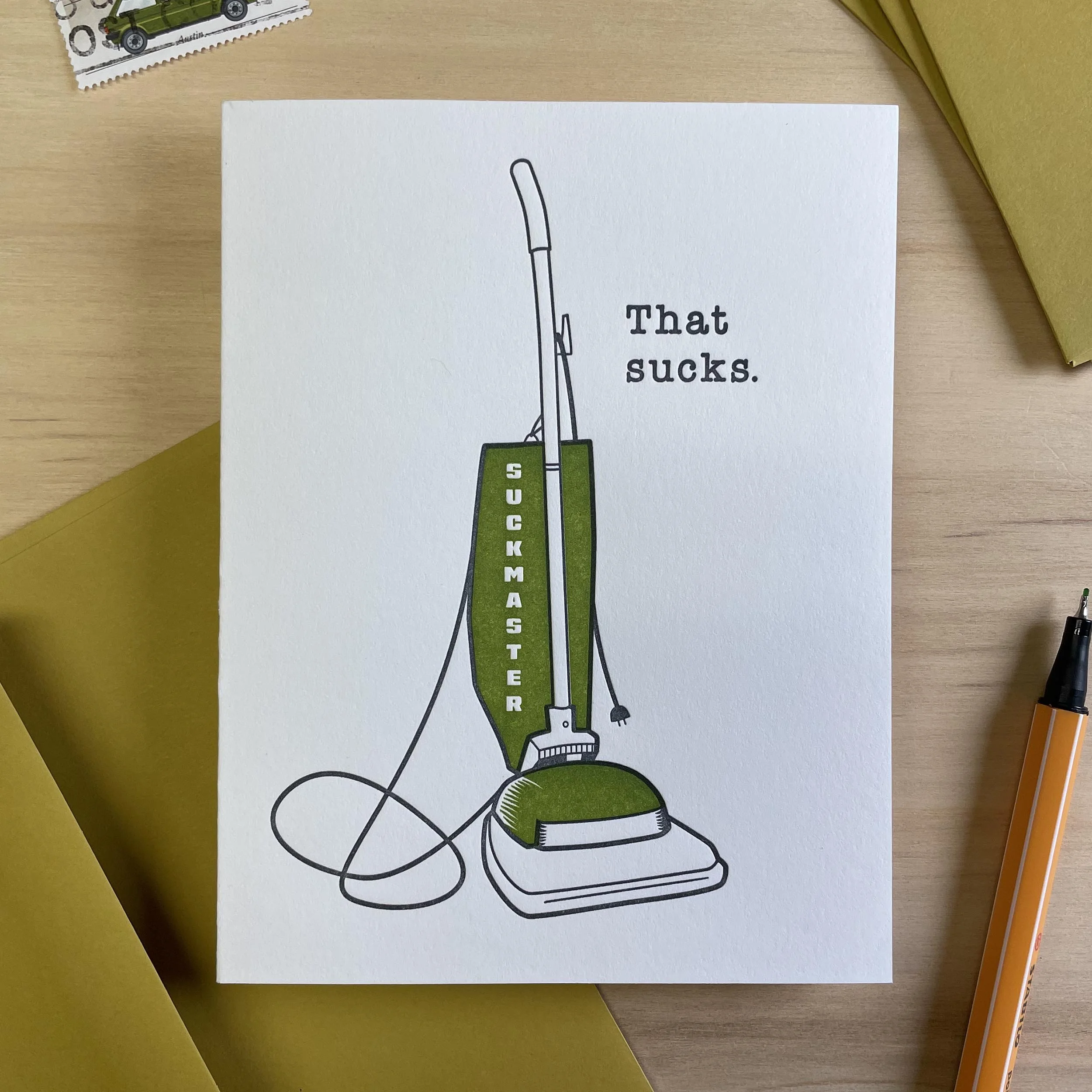 White letterpress card with a green and black drawing of a vintage upright vacuum. On the bag of the vacuum it says 'Suckmaster.' Next to the vacuum are the words 'That sucks.' Greenish gold envelope.