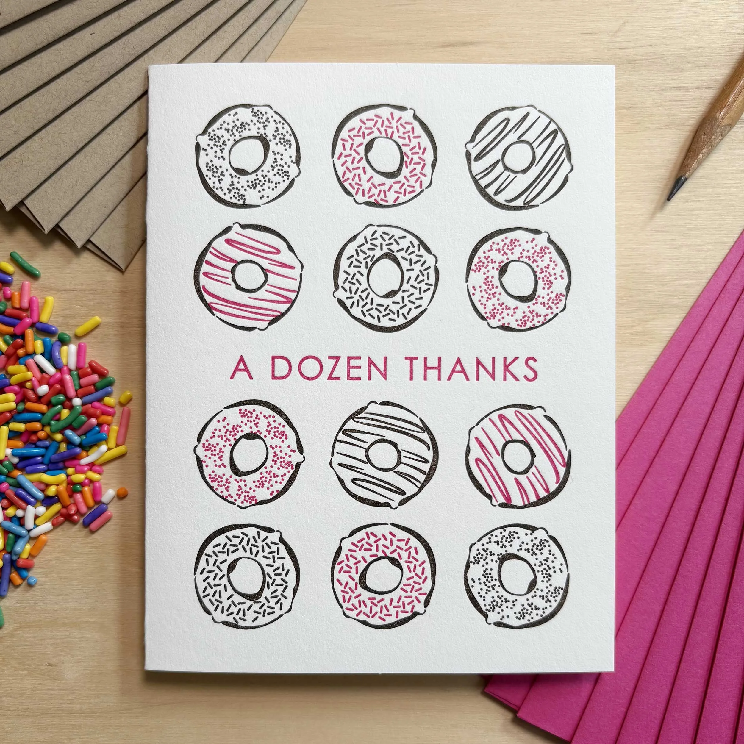 White letterpress card with 12 donuts printed in pink and dark brown. The text 'A DOZEN THANKS' is printed in pink in the center of the card. Pink and kraft envelopes.