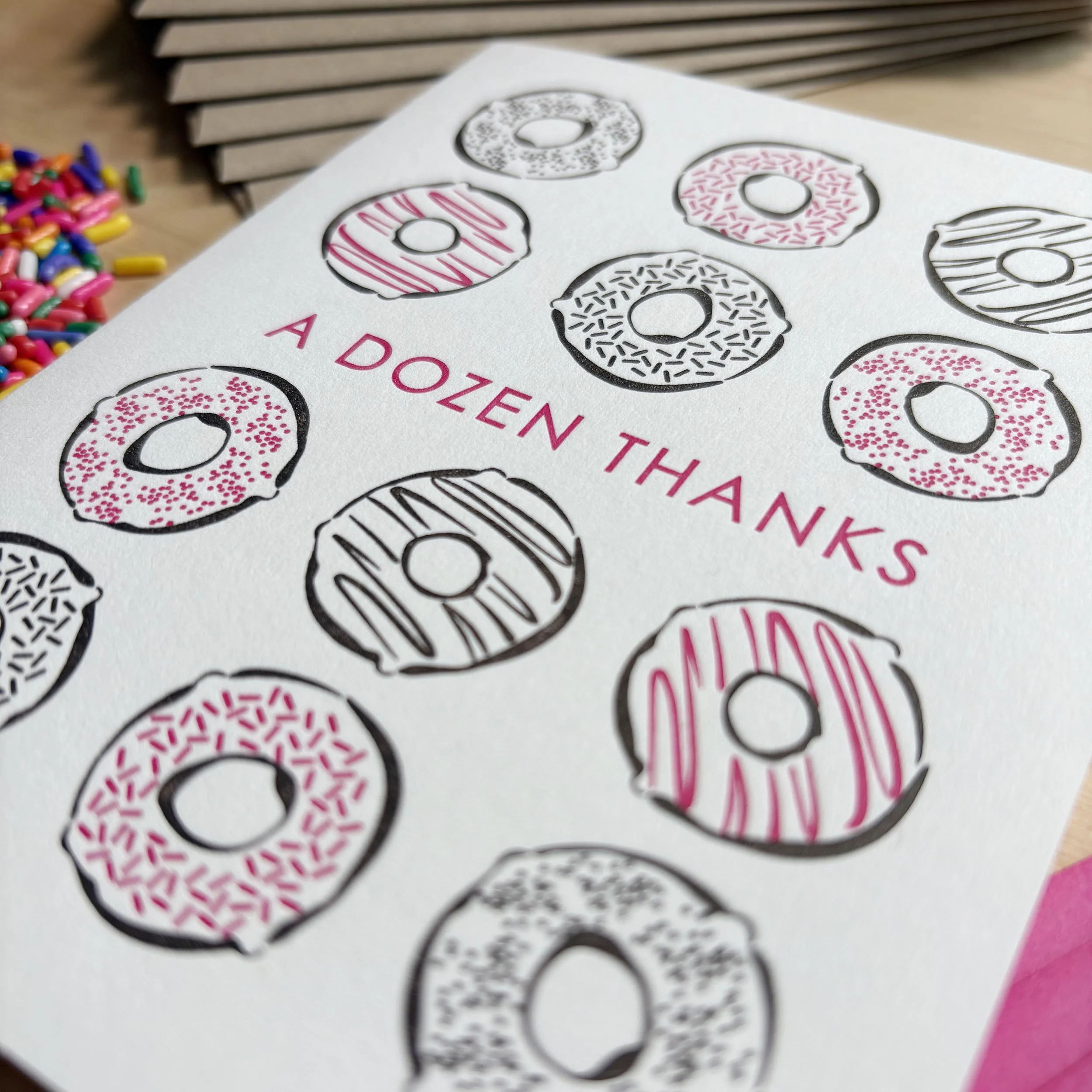 Close up of white letterpress card with 12 donuts printed in pink and dark brown. The text 'A DOZEN THANKS' is printed in pink in the center of the card.