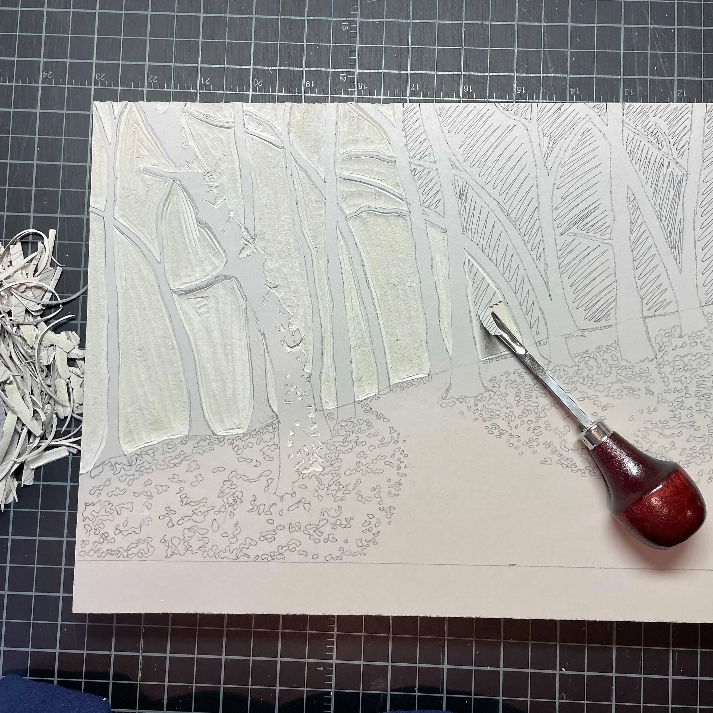 A light gray linoleum block is shown partially carved. The carving tool and some of the shavings are nearby. The image being carved is a woodland scene with leaves and a trail leading into the woods.