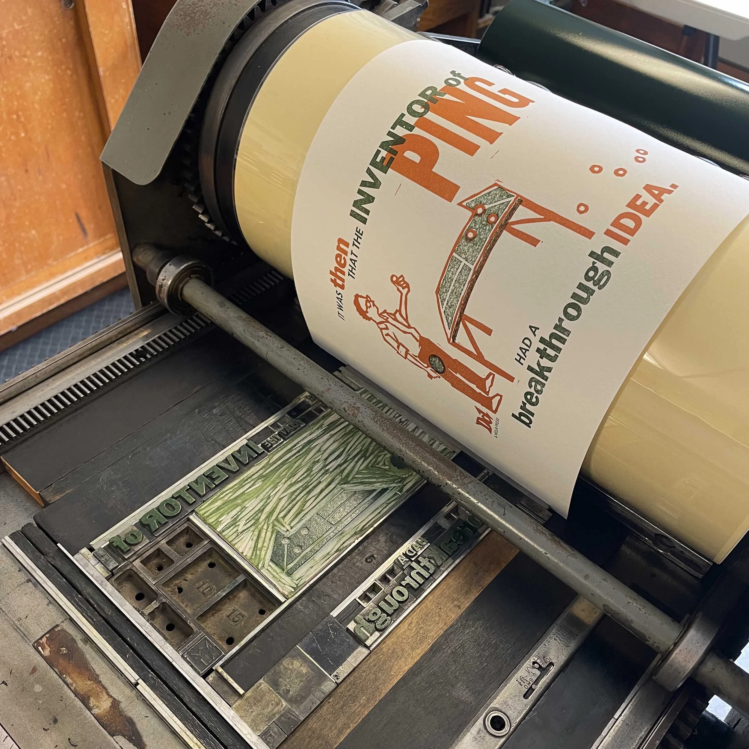 A vintage printing press printing green ink on white paper. The print features orange and green text that reads, 'It was then that the inventor of 'Ping' had a breakthrough idea.'