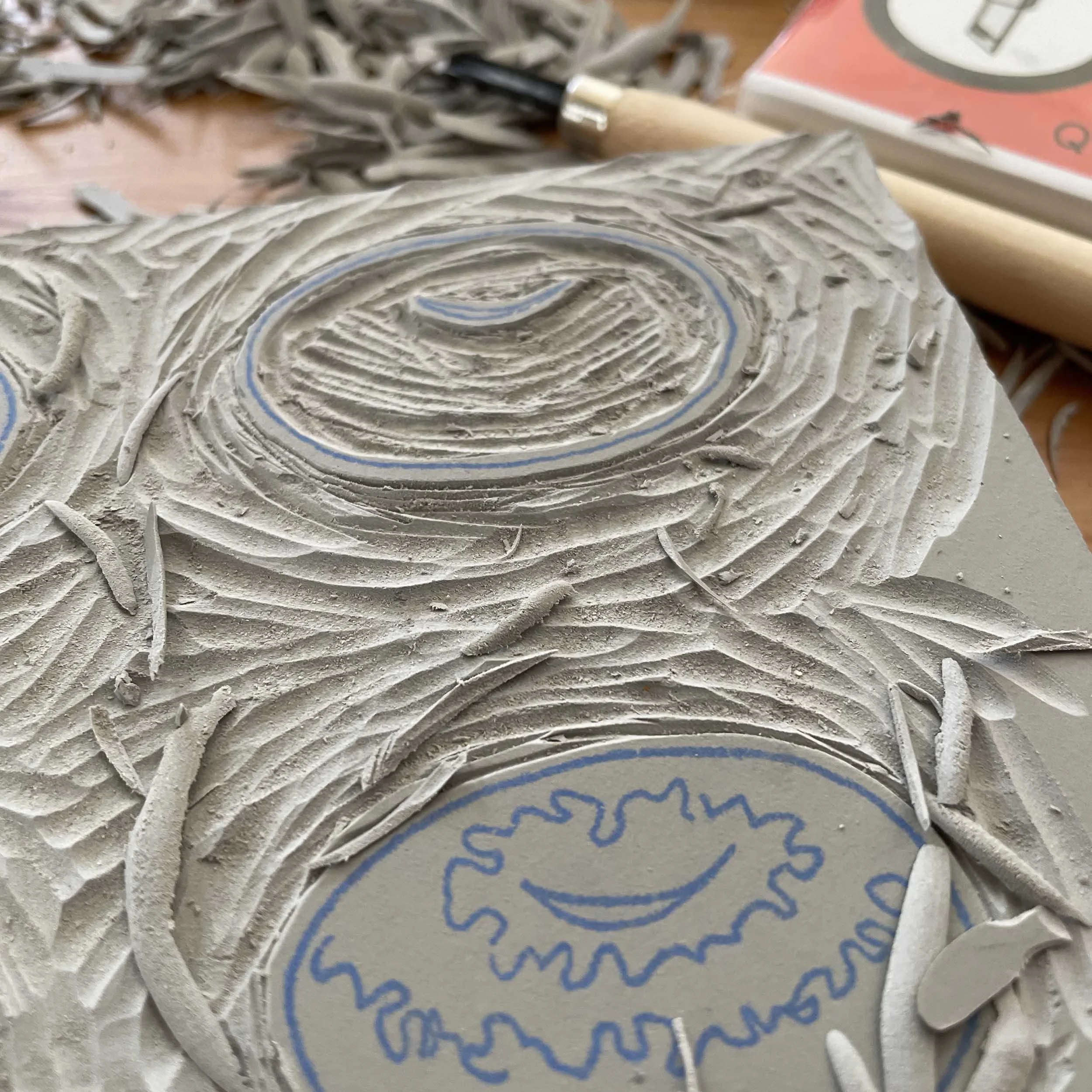 Carving of a linoleum block for Bagel Story by Drew Dernavich. The linoleum is gray with blue line drawings by Dernavich. Scraps of linoleum have been carved away. Carving tool nearby.