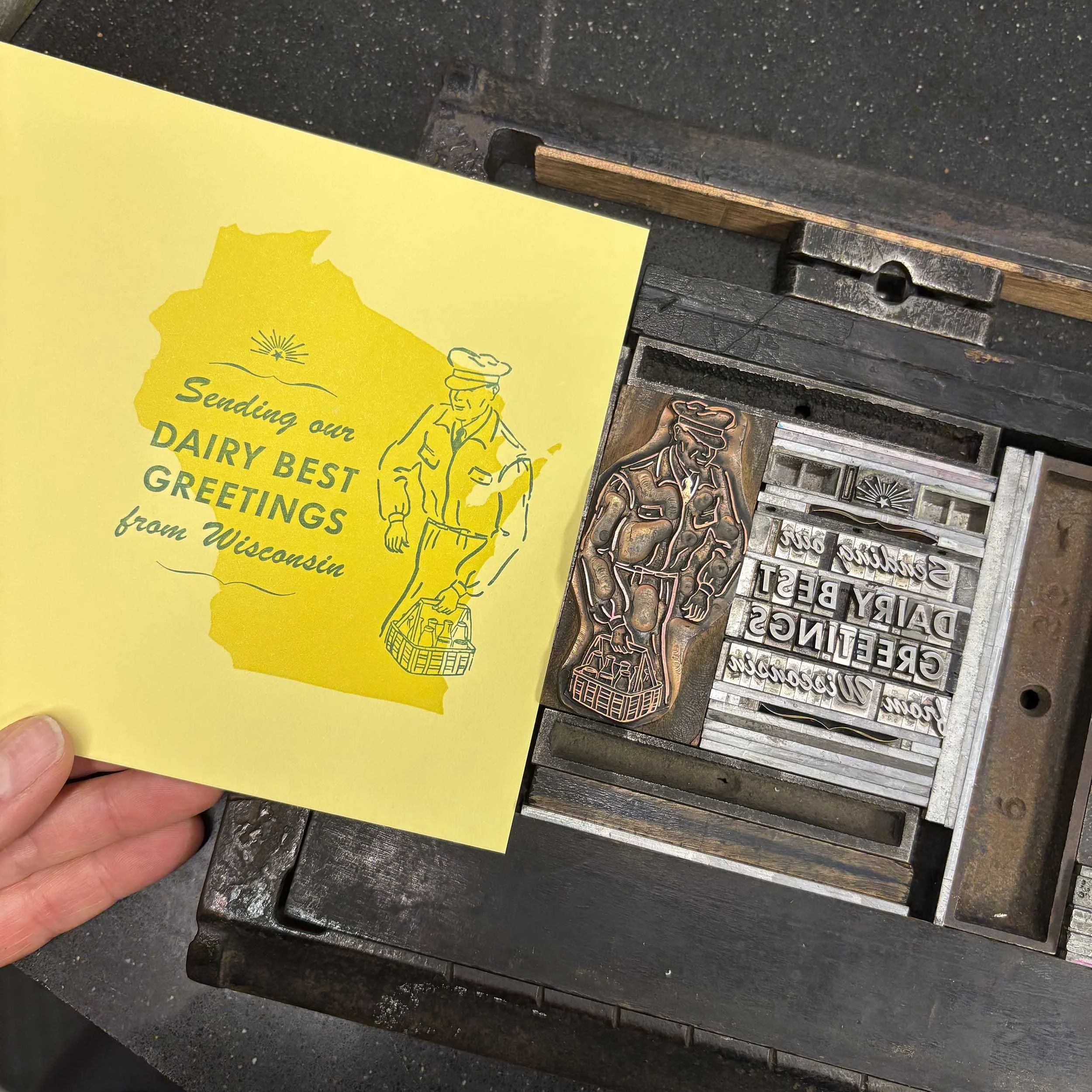 Letterpress lockup for a card with green type and a vintage milkman graphic printed over a solid yellow Wisconsin on light yellow paper.The words say 'Sending our dairy best greetings from Wisconsin.'