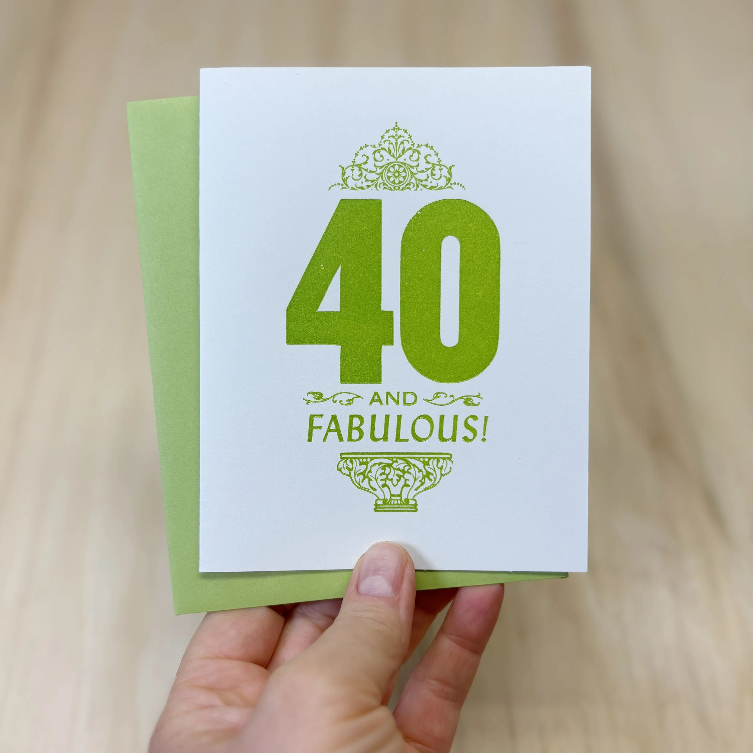 A hand is holding the 40 and Fabulous letterpress card. Bold 40 with smaller words AND FABULOUS. Letterpress ornaments add decoration. Green ink on white paper. Green envelope.