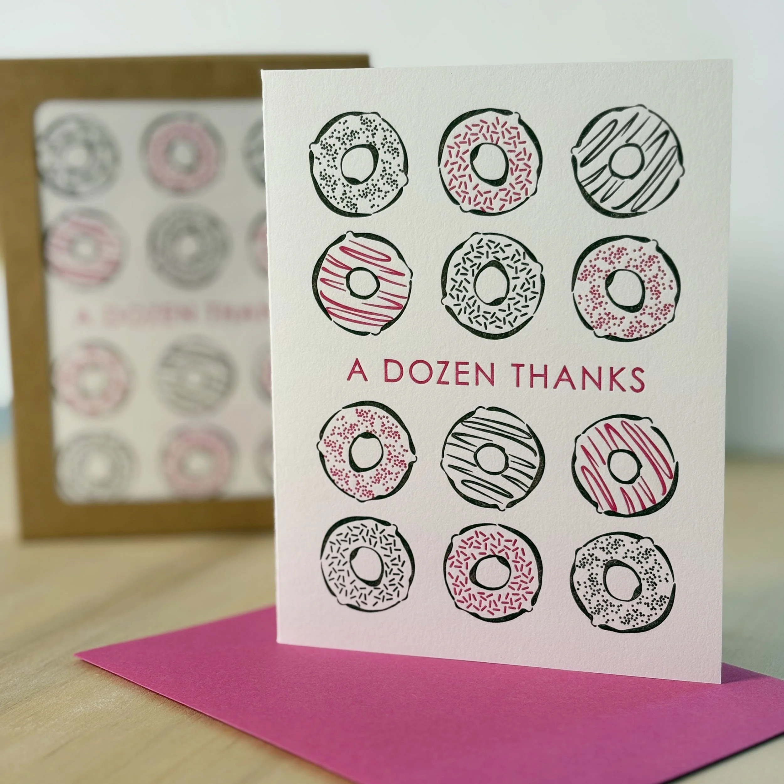 White letterpress card with 12 donuts printed in pink and dark brown. The text 'A DOZEN THANKS' is printed in pink in the center of the card. Pink envelope.