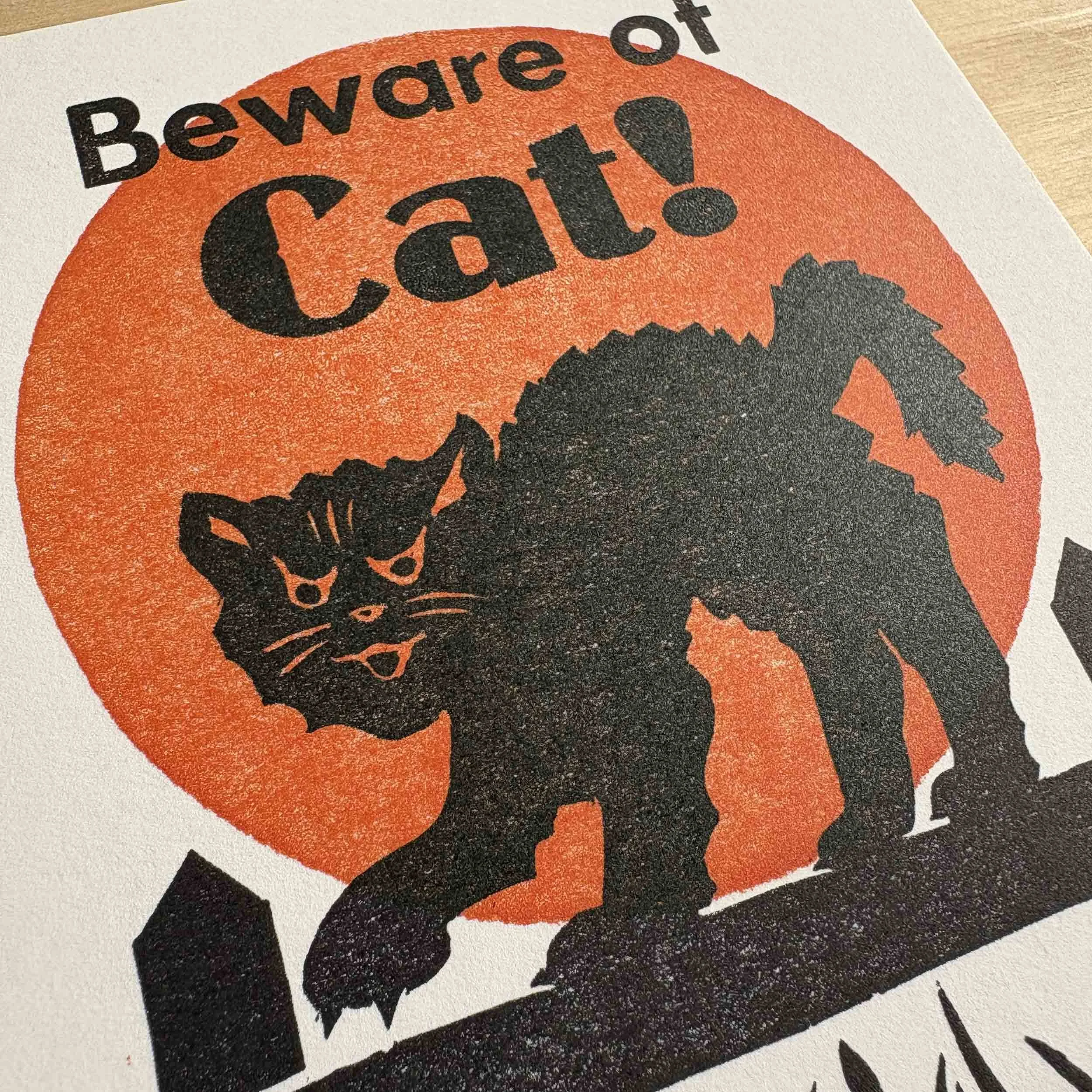 'Beware of Cat!' art print. Black text on white paper above a spooky black cat walking on a fence in front of an orange full moon.