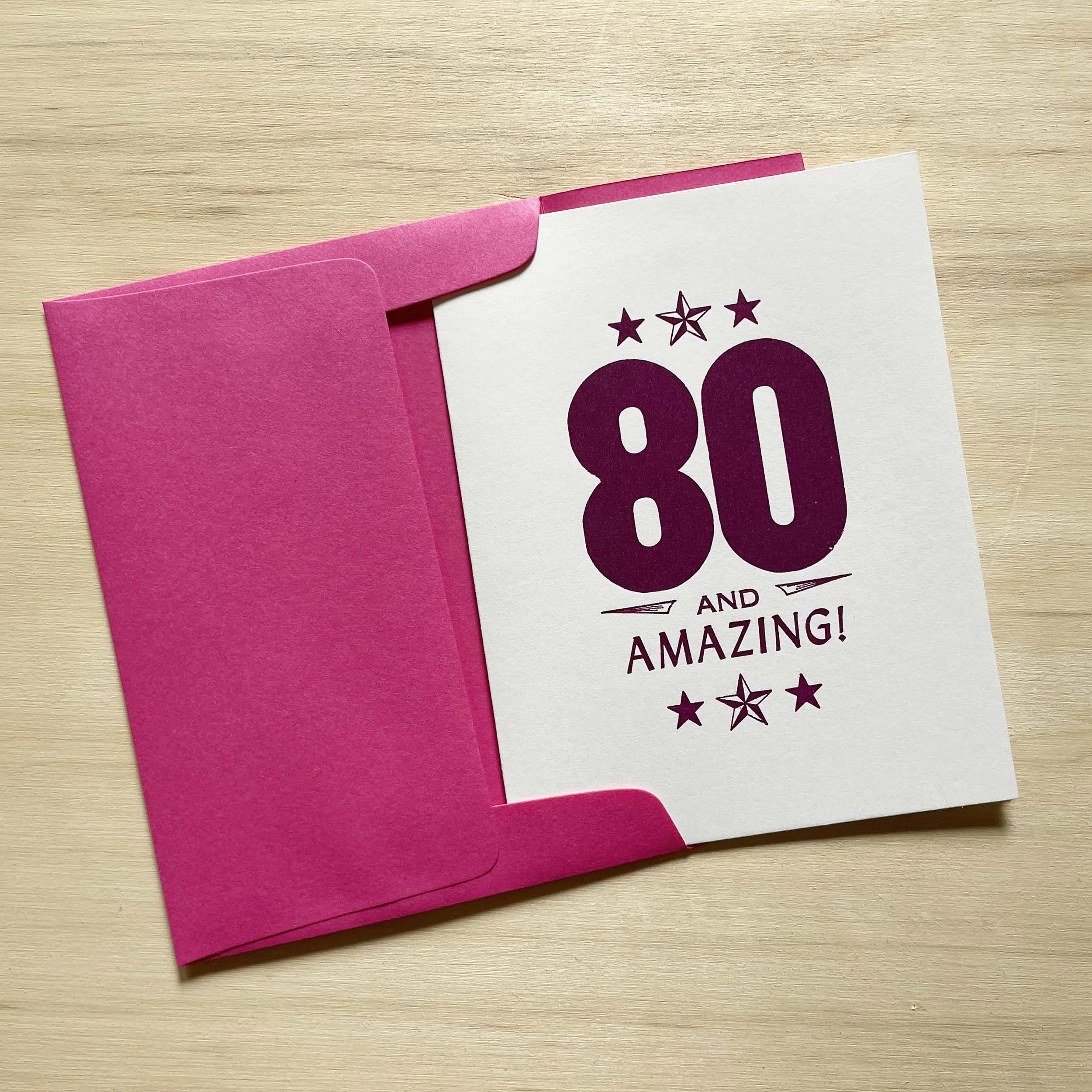 80 and Amazing card features a bold 80 with smaller words AND AMAZING. Letterpress ornaments add decoration. Dark purple ink on white paper. Magenta envelope.