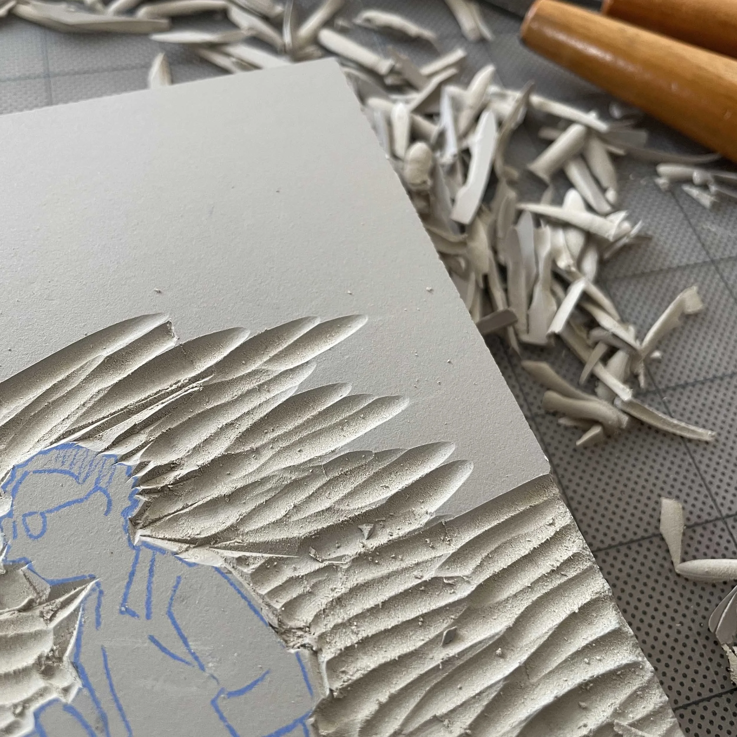 Close-up view of a linoleum block with carving in progress. A light gray block shows a partially carved design; a figure with glasses and a shirt with sleeves rolled up is sketched in light blue.