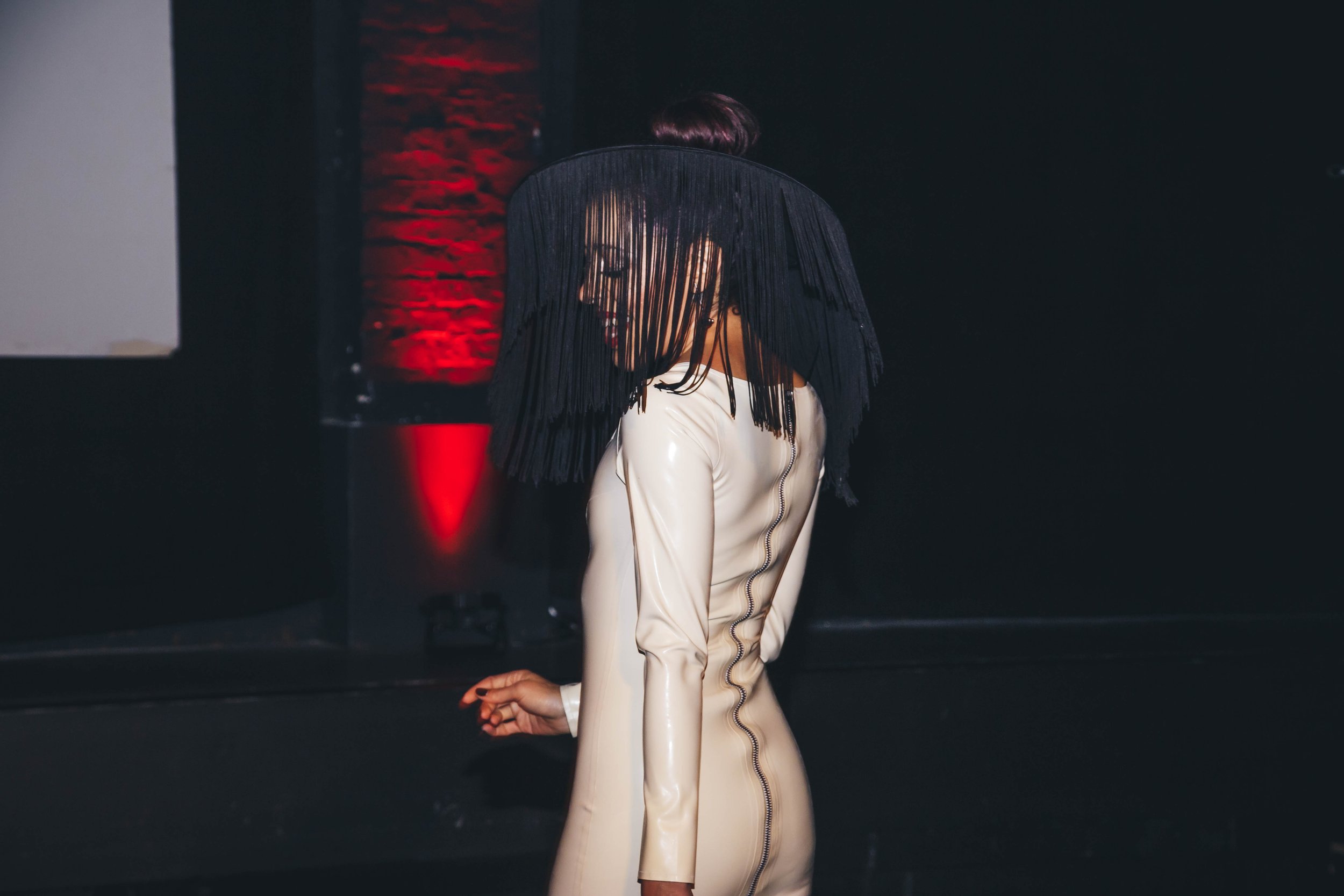 A woman in a cream-colored dress with a fringe veil covering her face, standing in a dark room with red lighting and a brick wall in the background.