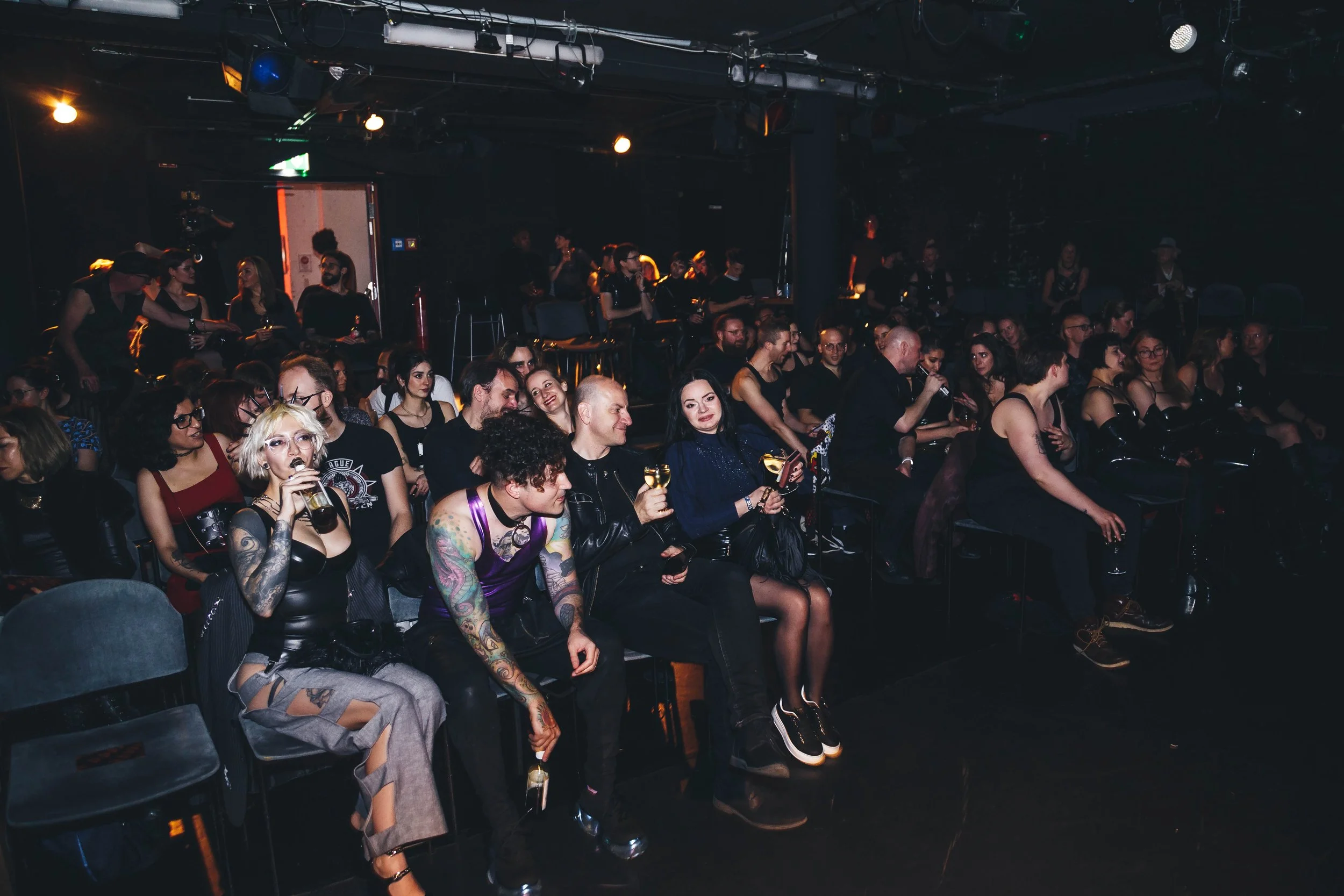 An audience at a concert or comedy show in a dark indoor venue, seated and enjoying drinks.