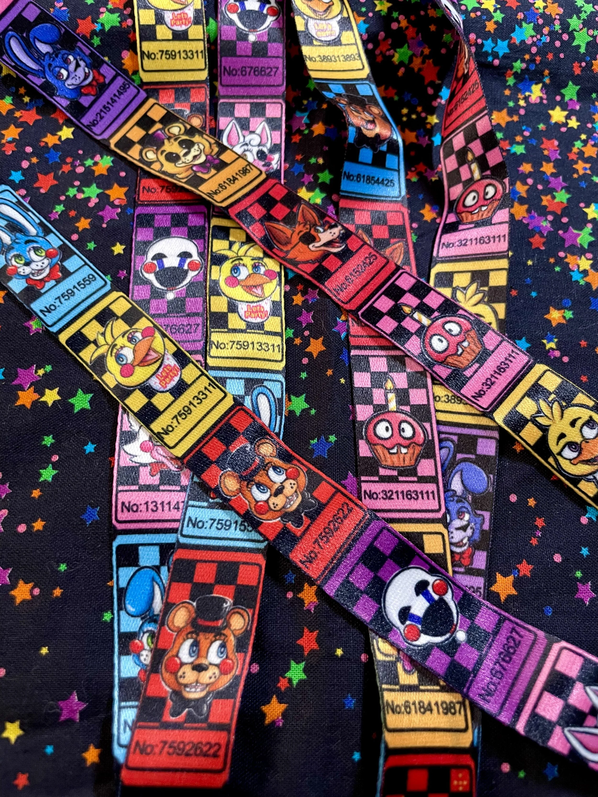 Colorful wristbands featuring characters from the video game franchise 'Five Nights at Freddy's,' with checkered patterns and serial numbers, laid over a starry background.