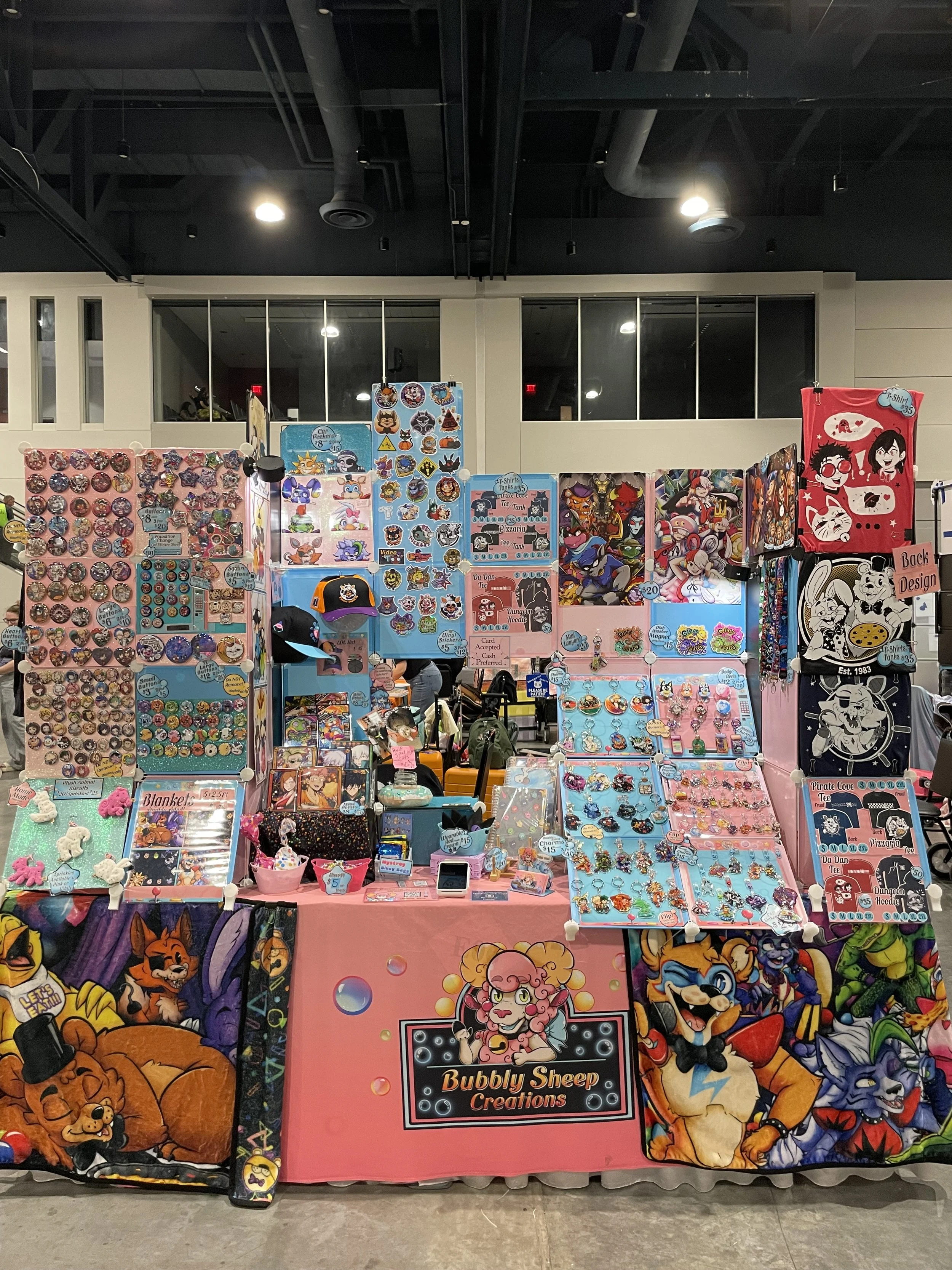 A colorful booth selling various pins, stickers, and accessories at an indoor event, with a sign reading "Bubbly Sheep Creations" and cartoon sheep and other animal illustrations on the table and backdrop.