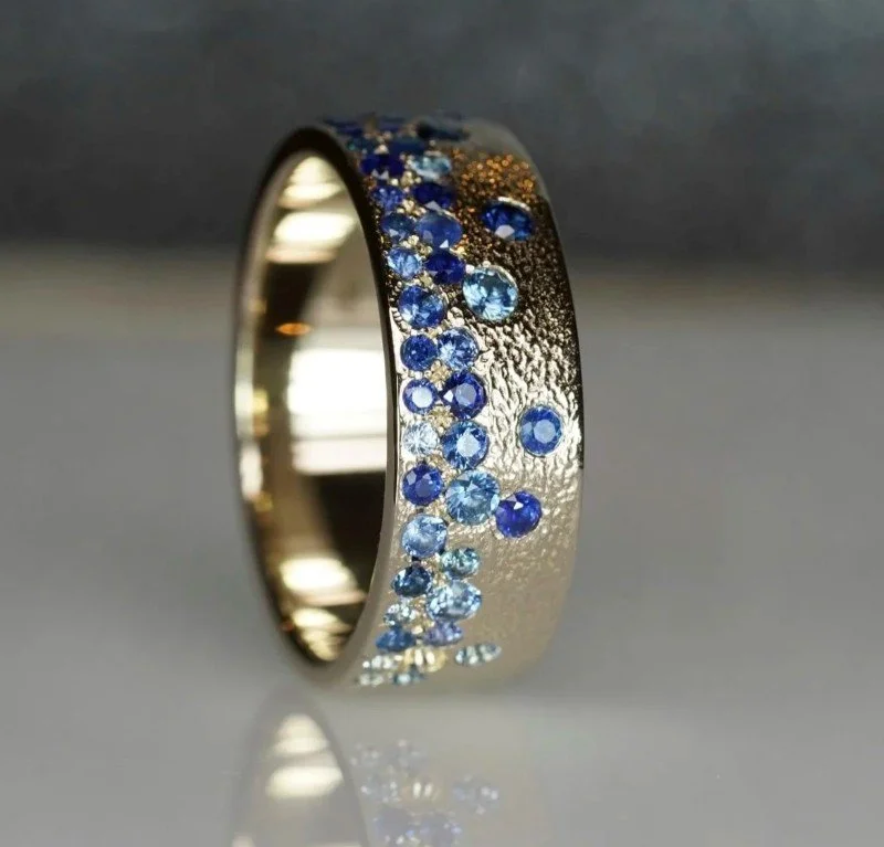 A wide gold band featuring a scattered cluster of various blue sapphires on a textured surface.
