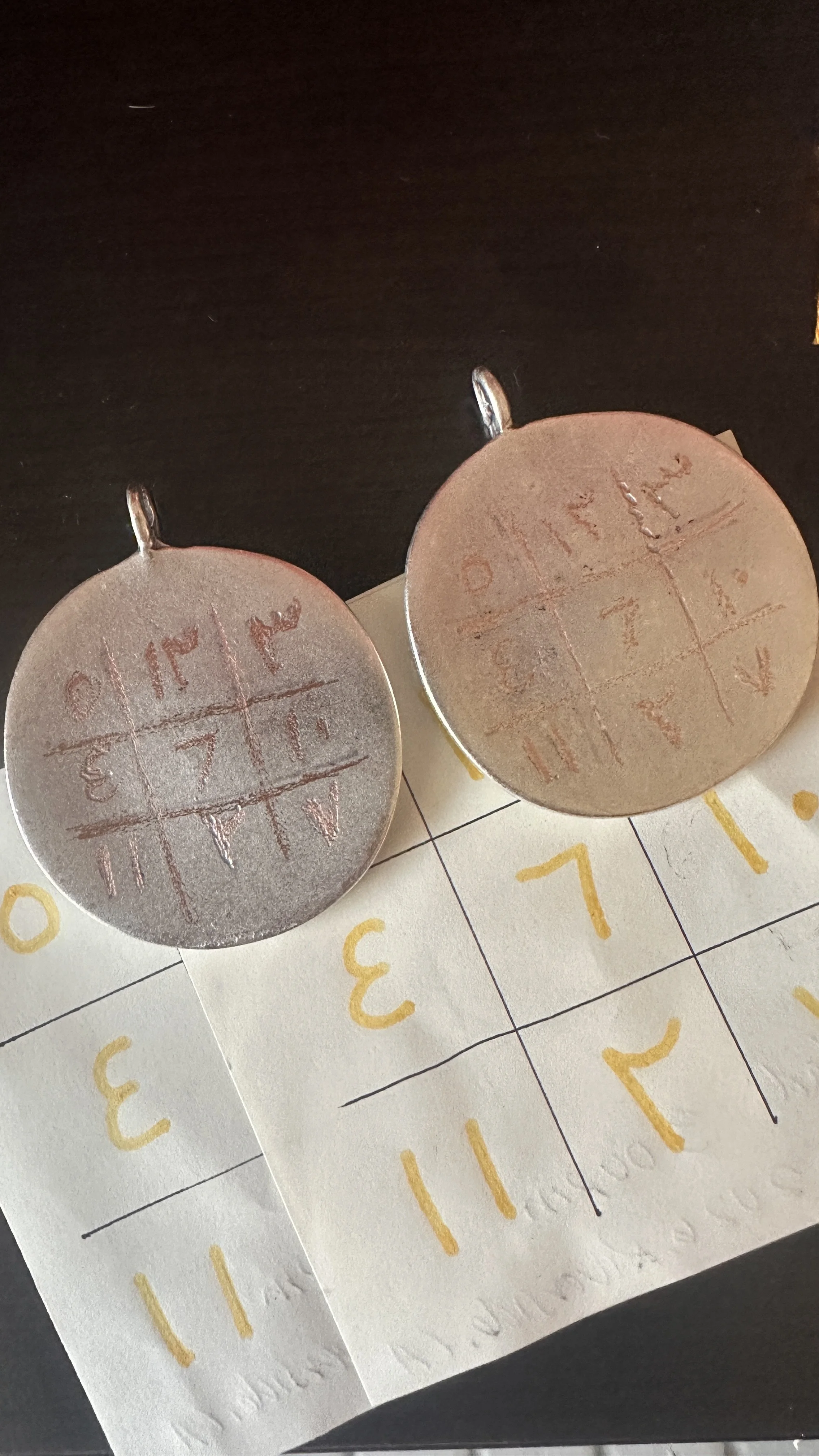 Two silver talismans with magic squares engraved on their surfaces, placed on a paper magic square using yellow-orange talismanic ink, on a dark surface.