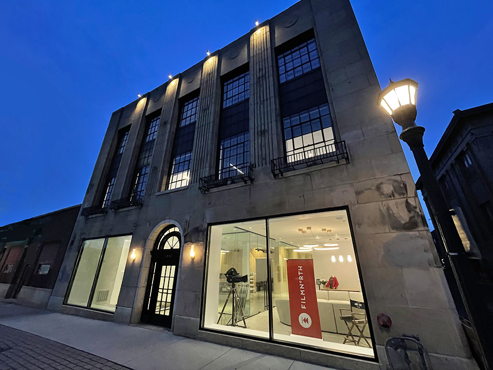 The stone Art Deco exterior of the FilmNorth building at dusk, with large windows showcasing a vintage film camera and a red branded banner inside