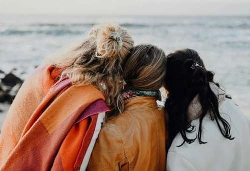 Three women standing close together on a beach, facing the ocean, with their heads touching, wearing casual jackets, with hair in various styles.