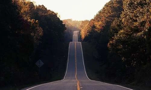 A long, winding road running through a wooded area with trees on both sides, with the road disappearing over a hill in the distance.