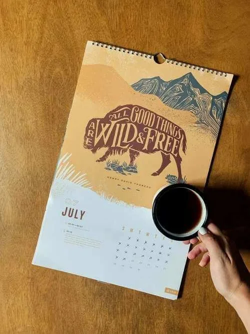 A wall calendar on a wooden surface, with a cup of tea held by a person's hand obscuring part of the calendar.
