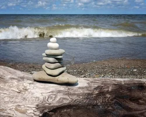 Stacked white and gray stones on a piece of driftwood on a beach with ocean waves and a partly cloudy sky in the background.