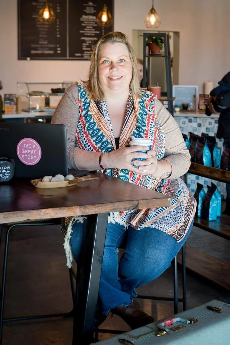 Smiling woman with blonde hair, wearing a colorful patterned top and jeans, sitting at a wooden table in a coffee shop, holding a mug, with a laptop nearby and warm lighting overhead.