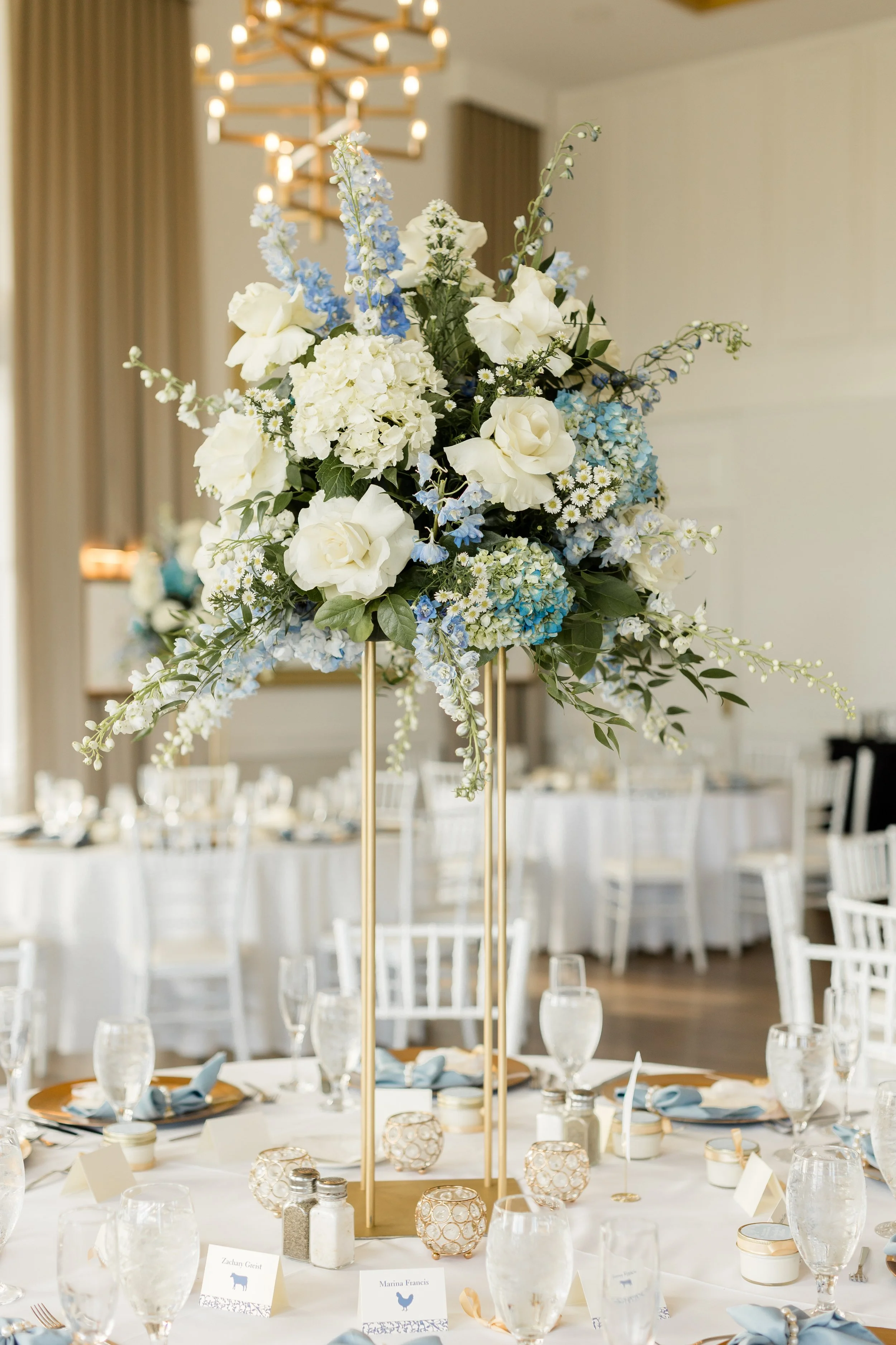 A tall floral centerpiece with white and blue flowers on a gold stand at a decorated banquet table.
