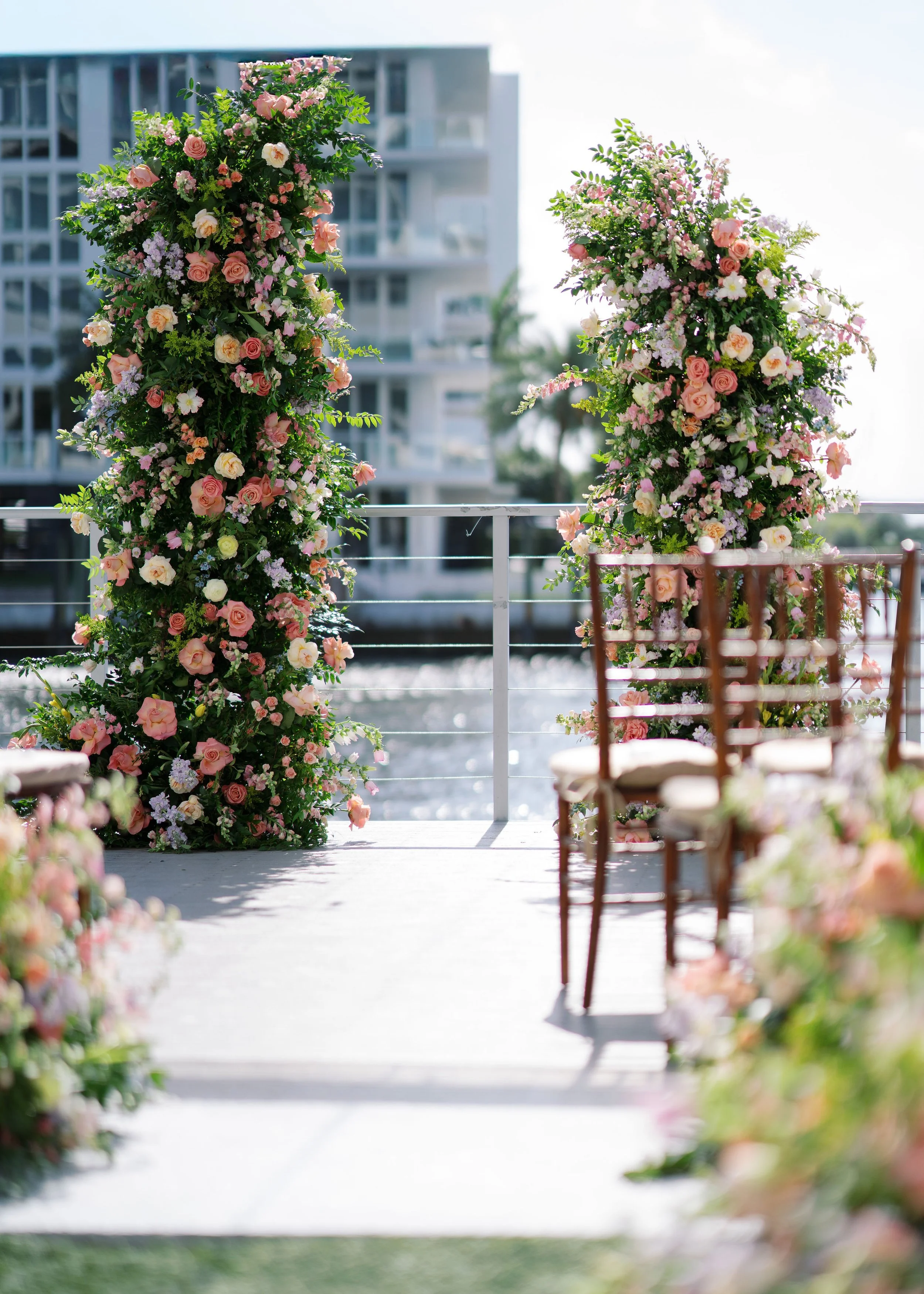 Two dimensional garden-style floral arch in pink, white, and peach roses and other flowers on a balcony overlooking water, with modern buildings in the background at a Fort Lauderdale wedding venue.