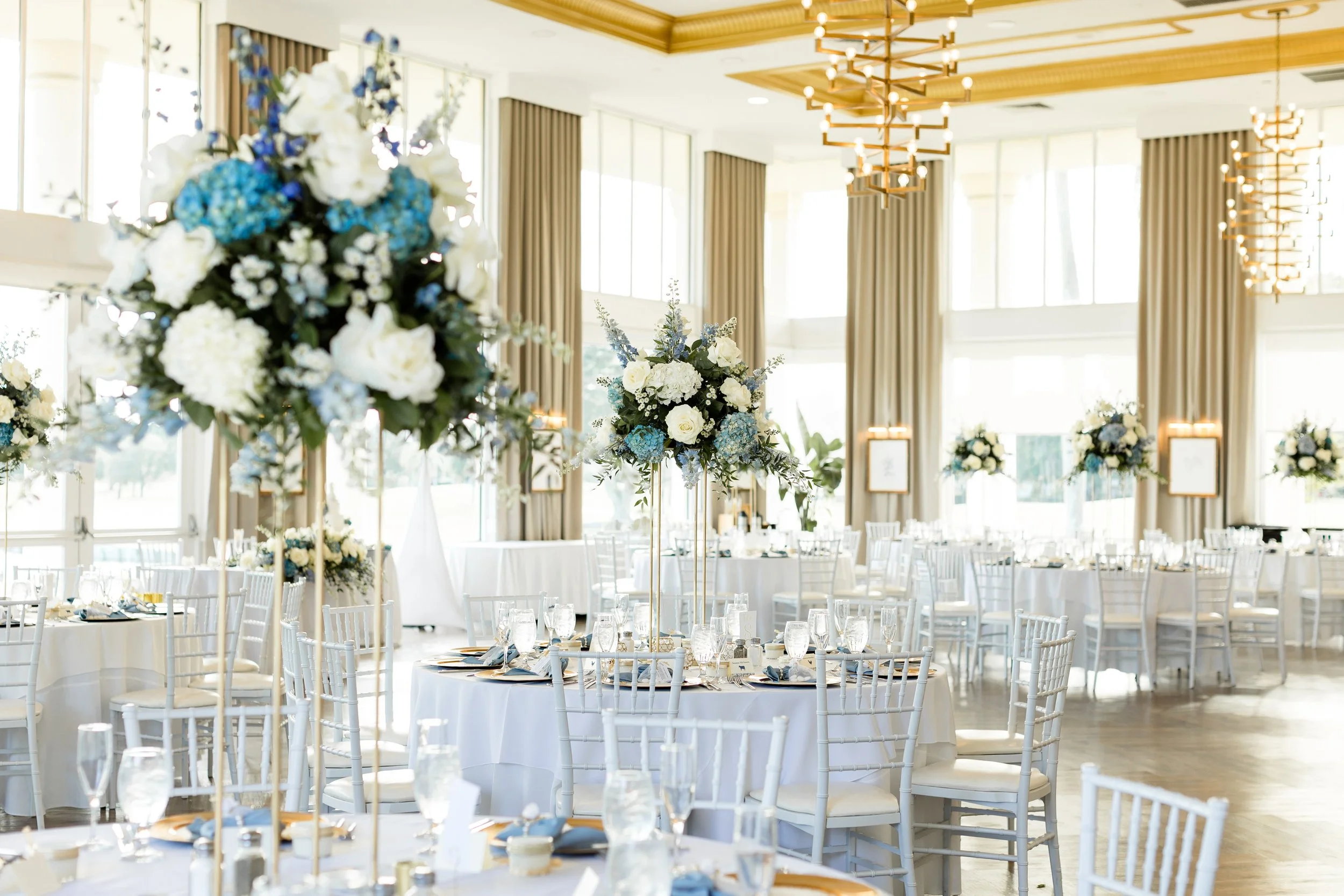 Elegant event decor with tall floral centerpieces featuring white and blue flowers, set in a bright, spacious room with large windows, framed by beige curtains, and surrounded by white chairs and tables with glassware and table settings.