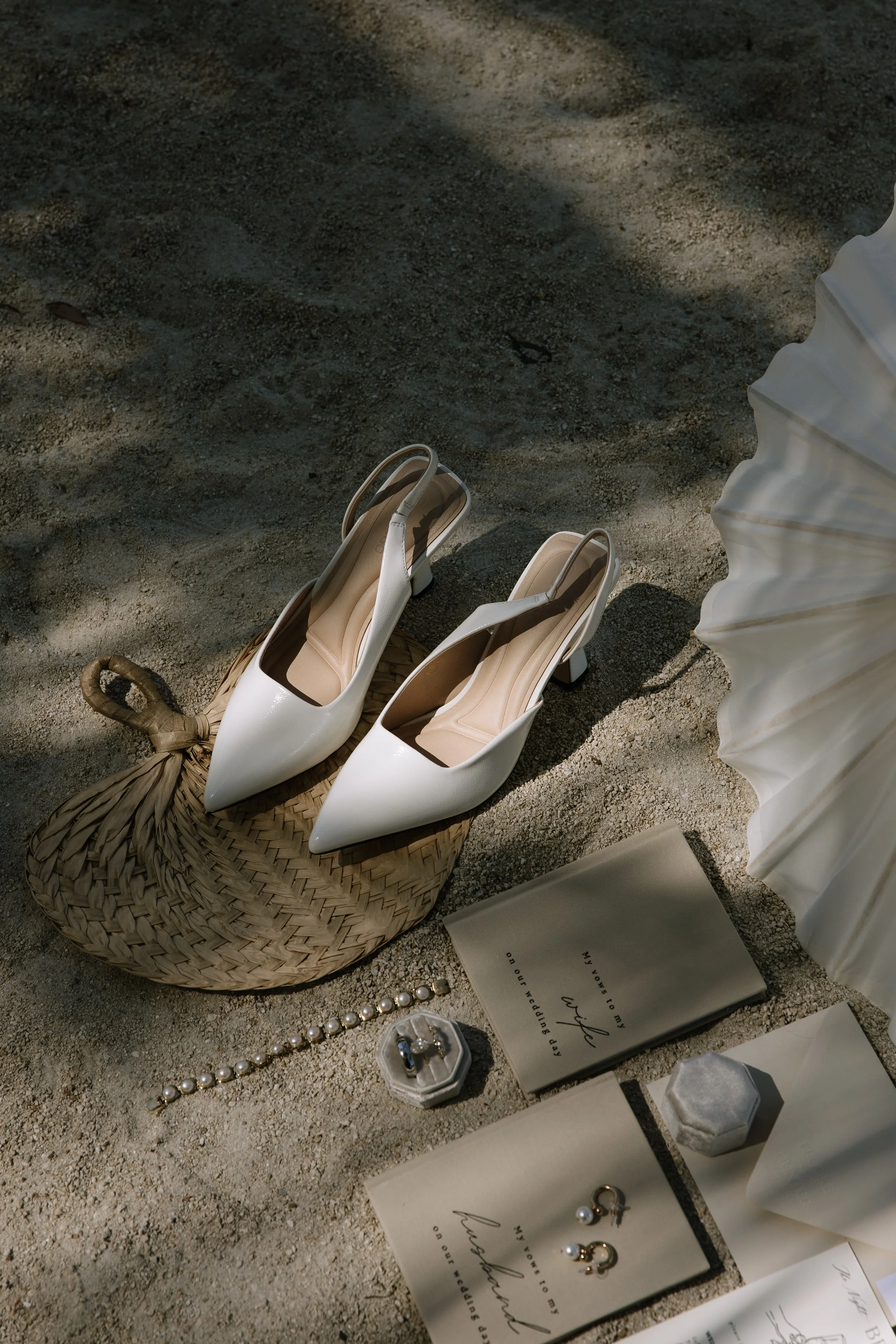 A pair of white high-heeled shoes placed on a woven basket, placed on sandy ground near a large white umbrella. Nearby are pearl jewelry and a book, with some jewelry also displayed on paper.