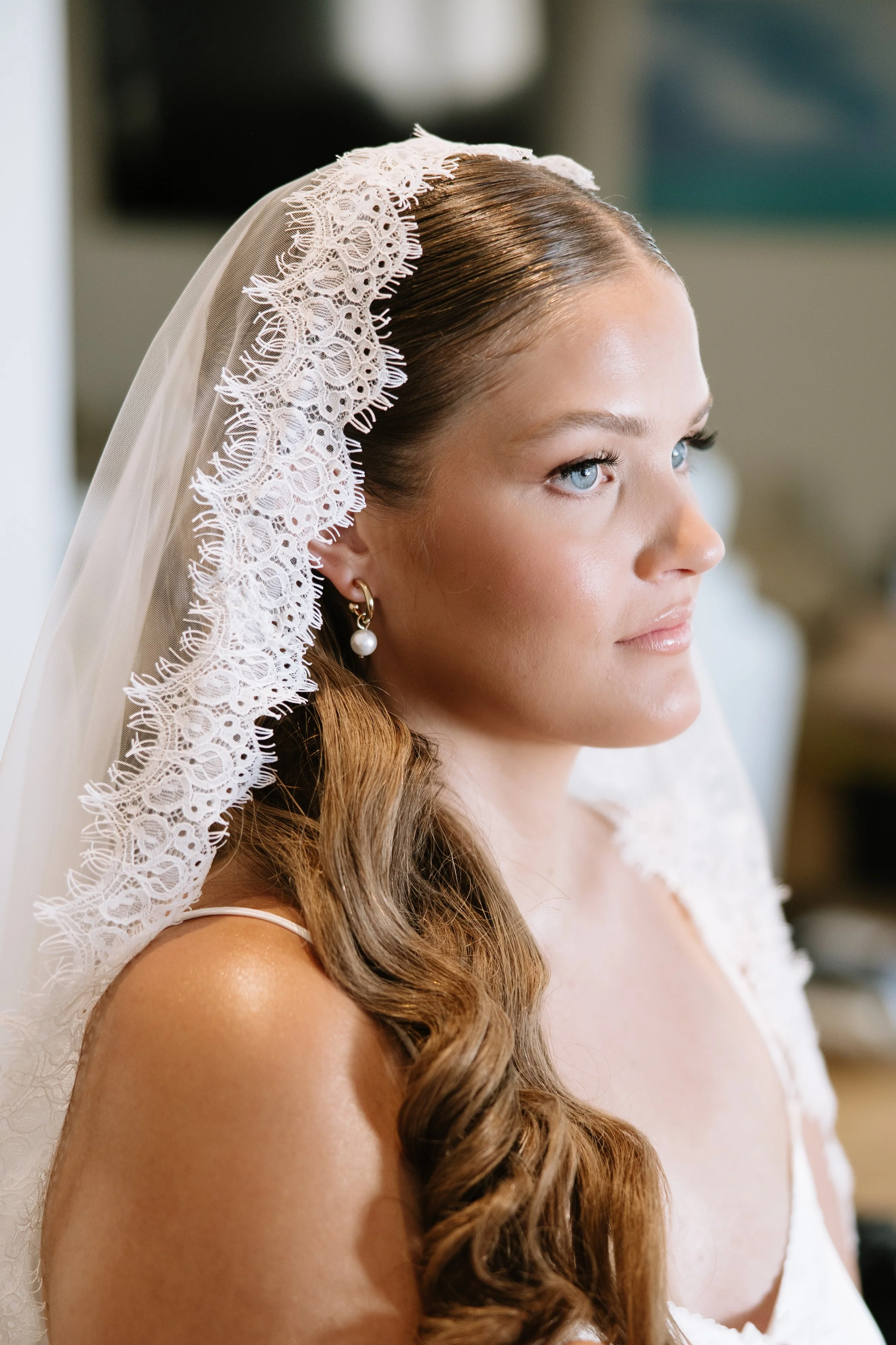 Close-up of a bride with long, wavy hair wearing a lace veil and pearl earrings, with soft natural makeup and a calm, confident expression.