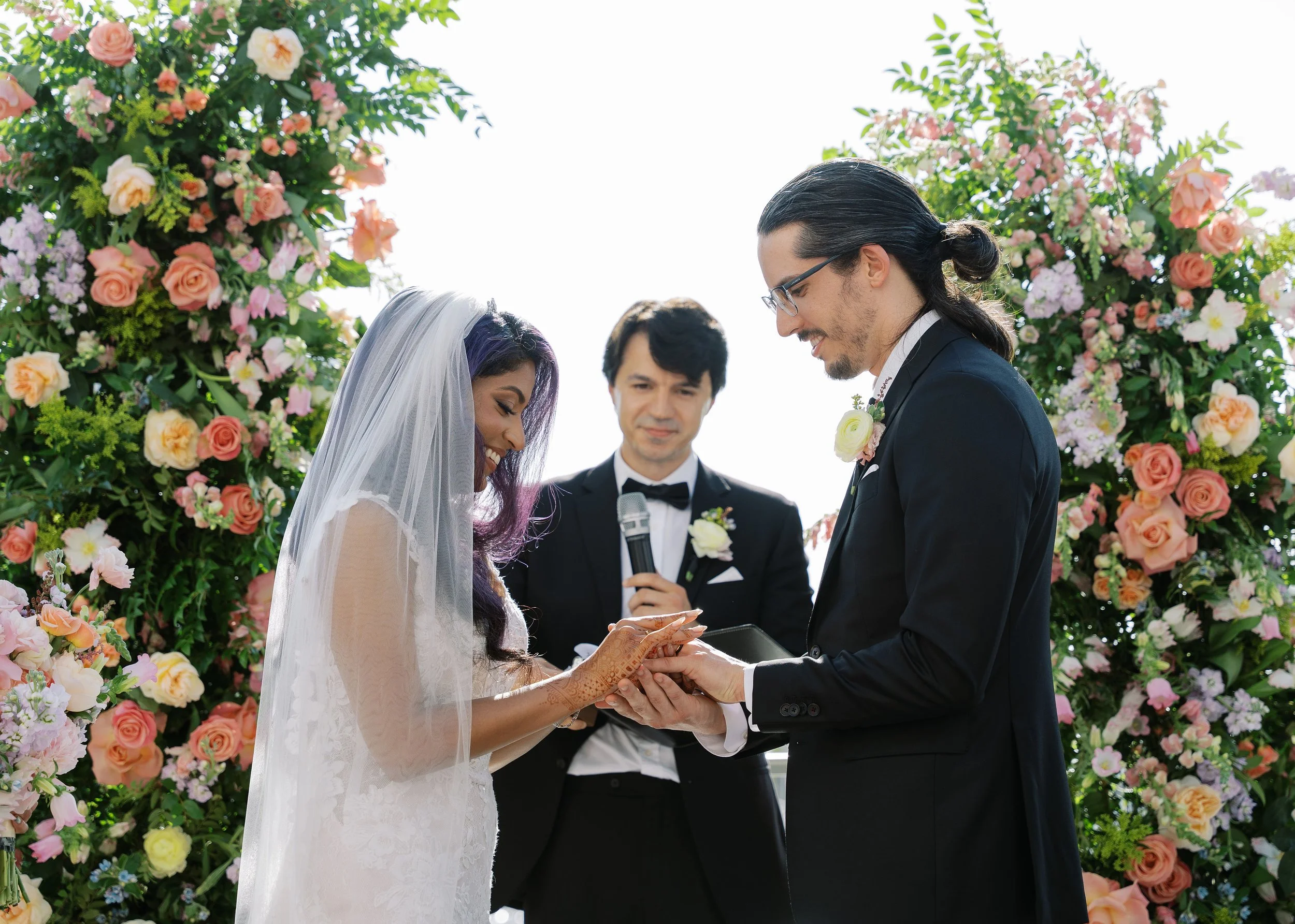 A bride and groom exchanging wedding vows outdoors at a ceremony decorated with lush floral arrangements, with a officiant overseeing the event.