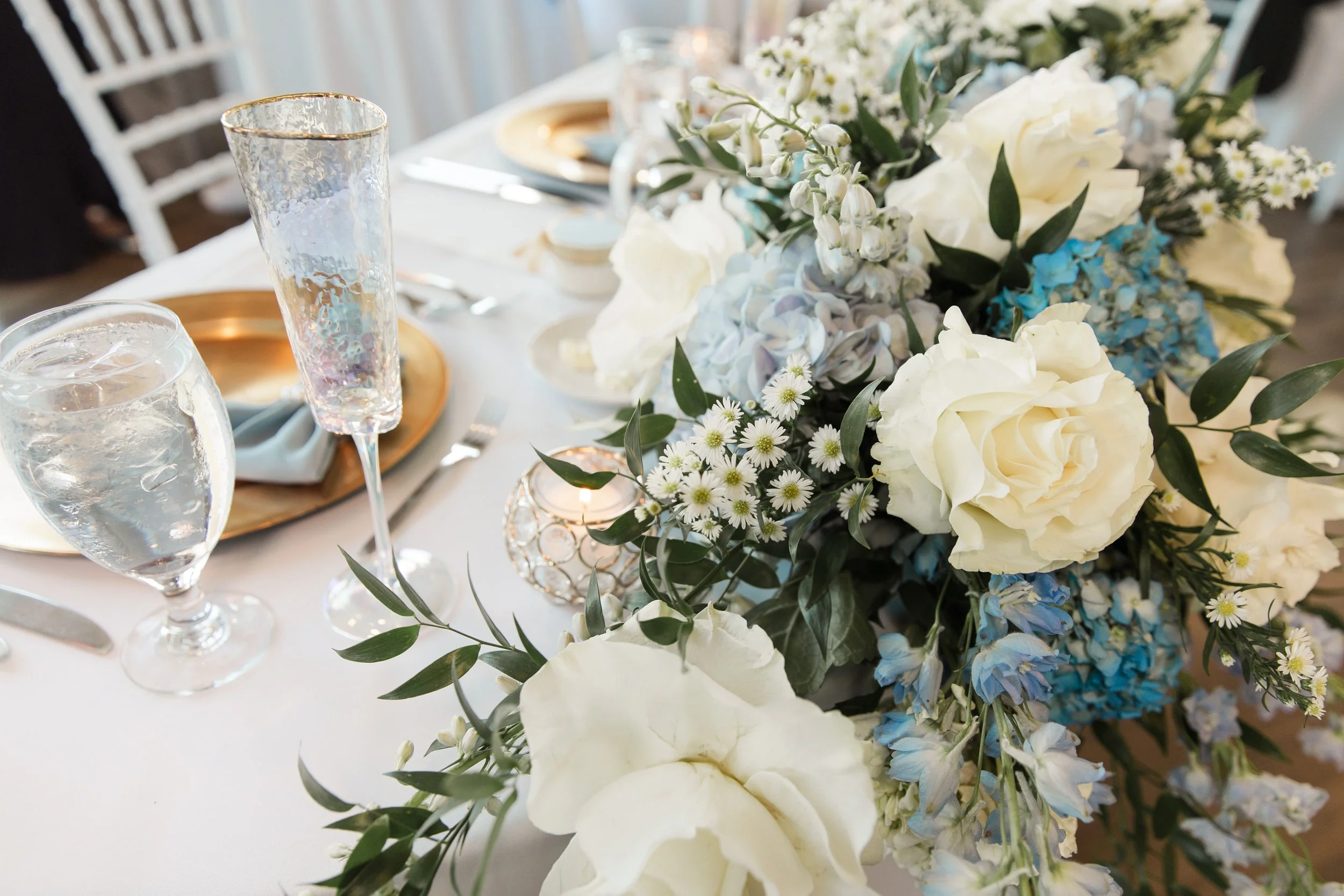 A close-up of a wedding table centerpiece with white and blue flowers, including roses, daisies, and hydrangeas, surrounded by greenery. The table is decorated with gold-rimmed plates, cutlery, glasses filled with water, and a small candle holder.