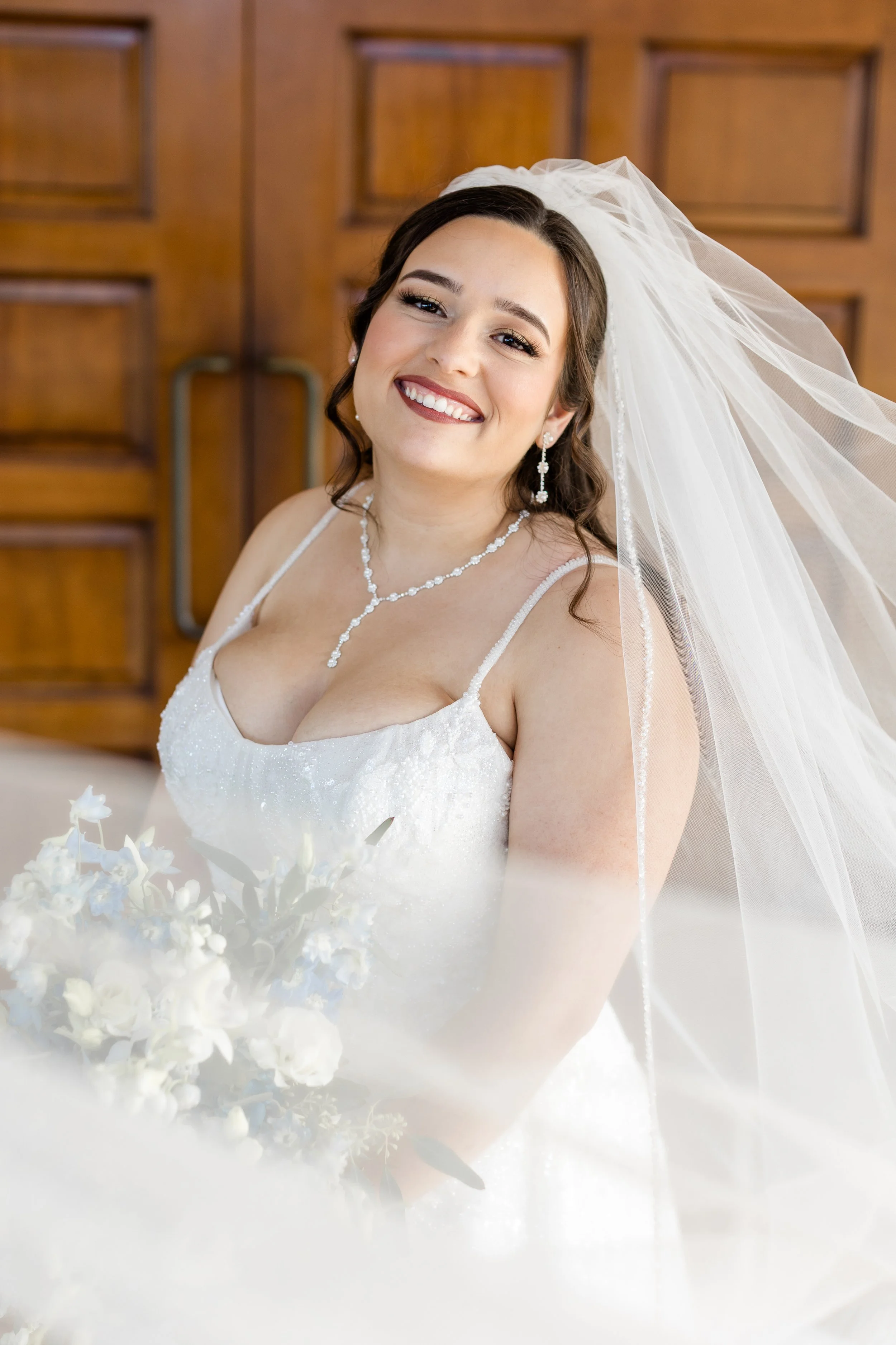A smiling bride in a white wedding dress with a veil and jewelry, holding a bouquet of white flowers, standing indoors with wooden doors in the background.