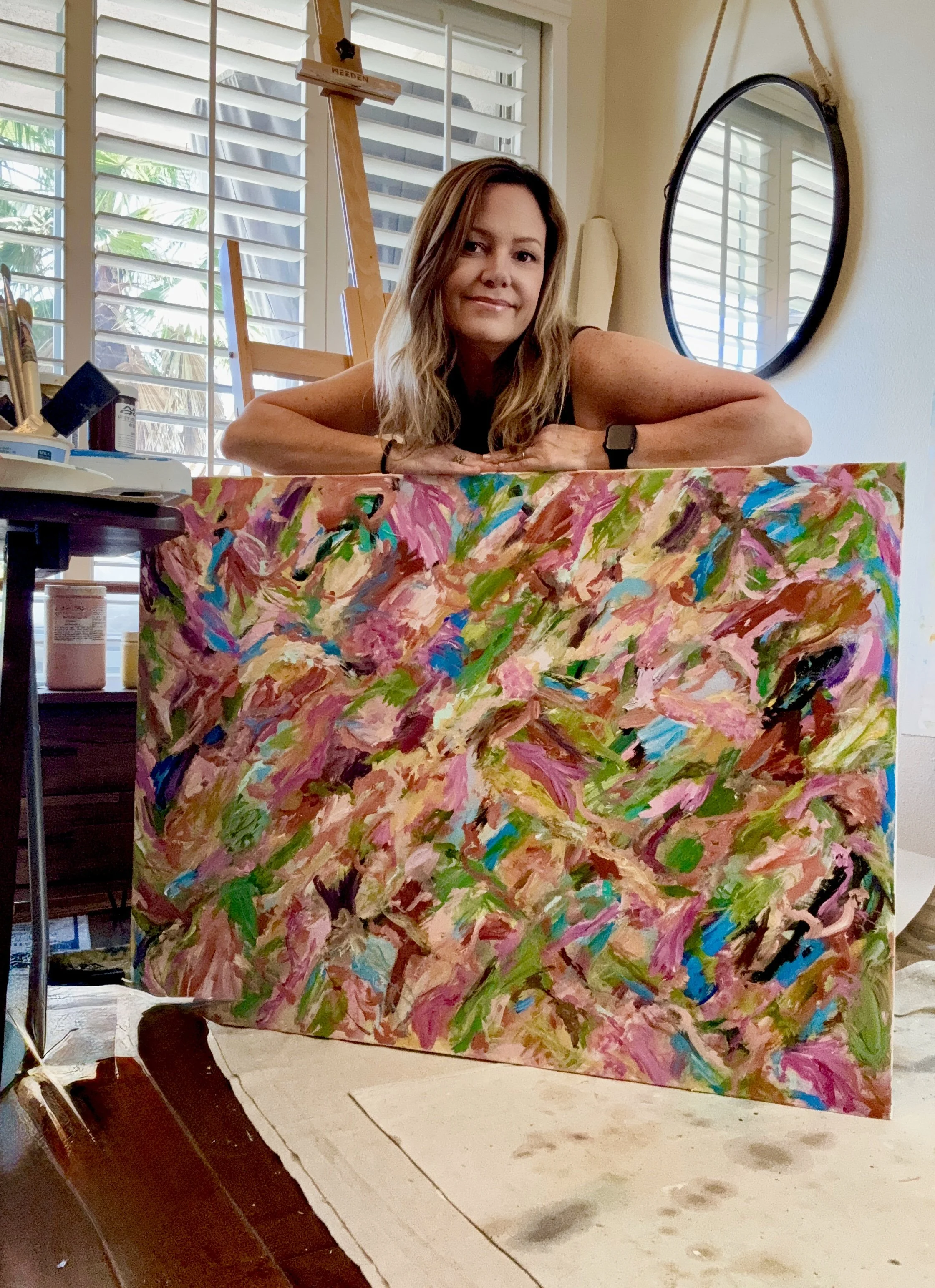 A woman with shoulder-length brown hair leaning on a colorful abstract painting in a room with large windows and shutters, side mirror hanging on the wall, office supplies on a desk, and an easel in the background.