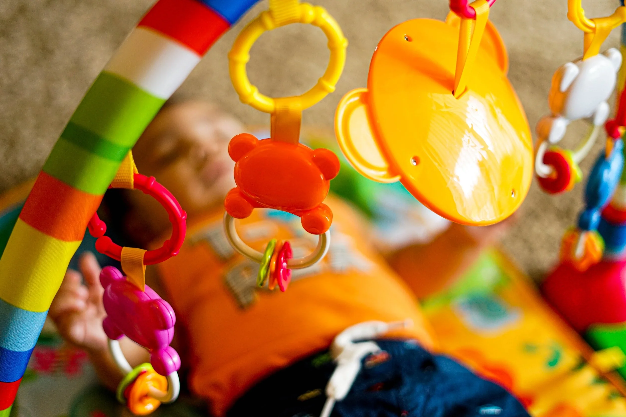 Child lying on the carpet playing with a colorful hanging toy featuring animals like a bear and a butterfly.
