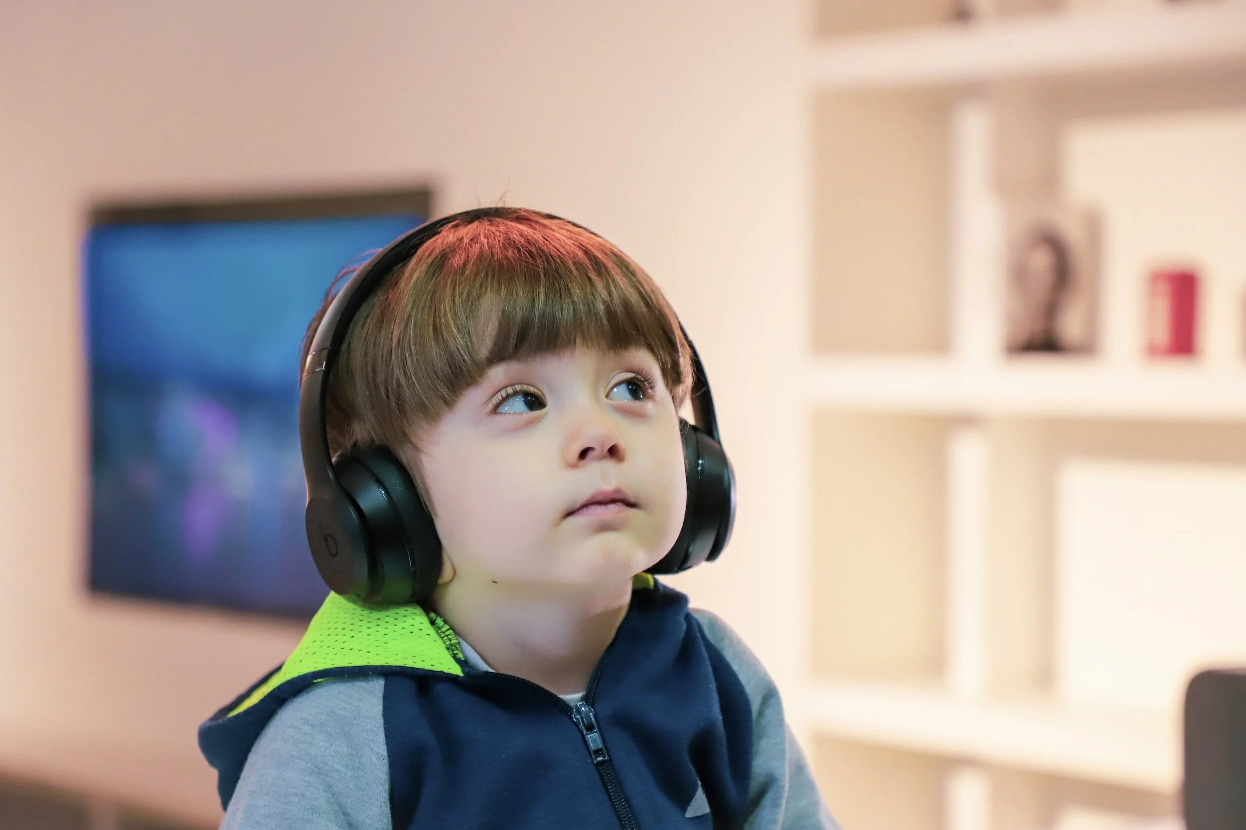 Young boy with brown hair wearing black over-ear headphones and a navy and gray hoodie, sitting indoors in front of a blurred background.
