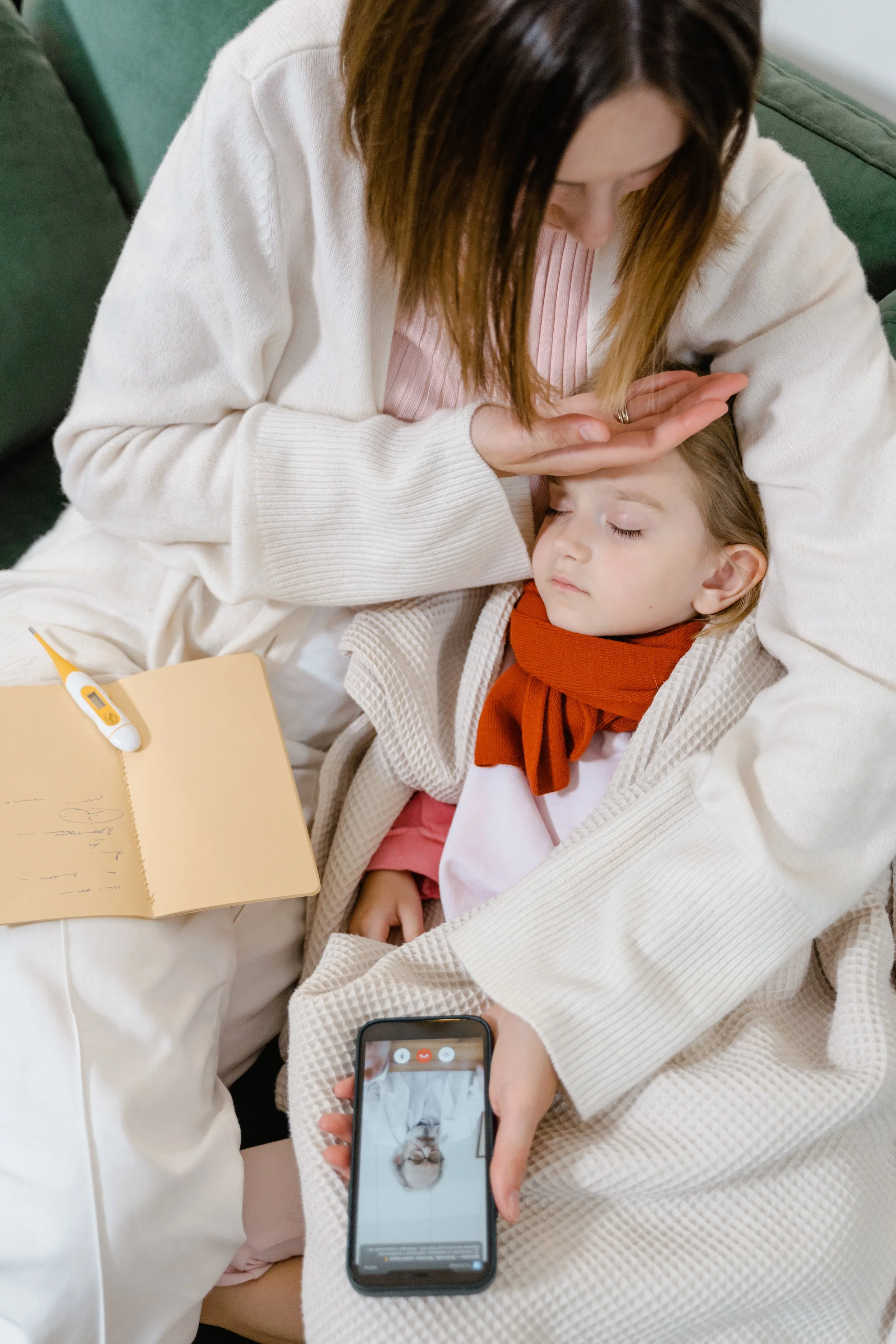 A woman tending to a young girl who appears to be ill, with a thermometer and notebook nearby, using a phone to record or call, indoors with a green sofa in the background.