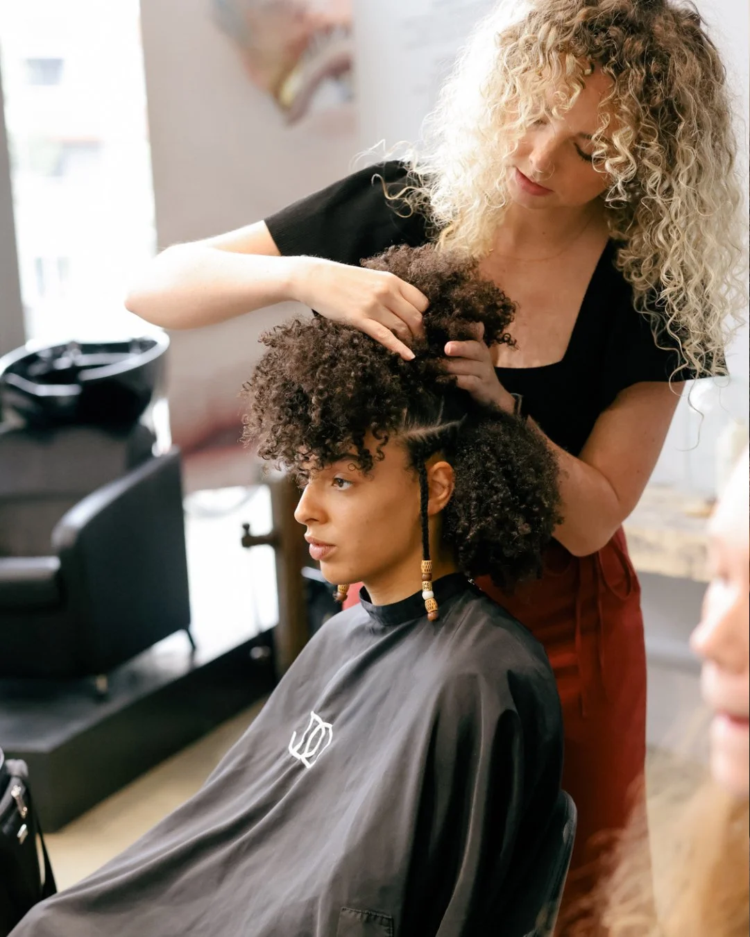 A hairstylist is styling a woman's curly hair in a salon. The woman is seated, wearing a black salon cape, and has beads in her braided sections of hair. The stylist, with blonde curly hair, is gently working on the client's hair.