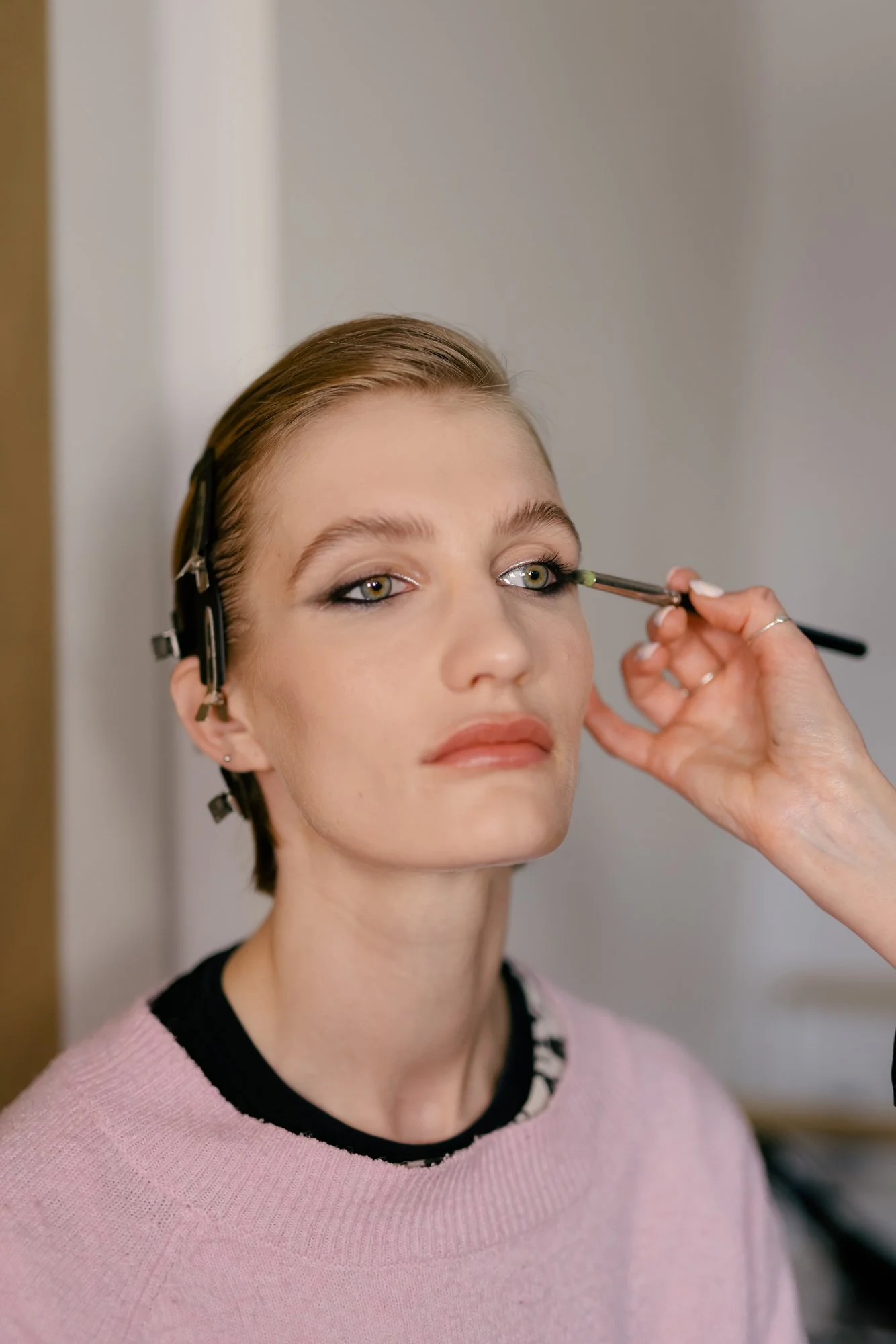 A woman with short brown hair and light skin getting her eye makeup applied by a makeup artist. The makeup artist is using a brush near her right eye, applying eyeshadow or eyeliner. The woman is wearing a pink sweater and appears calm and focused.