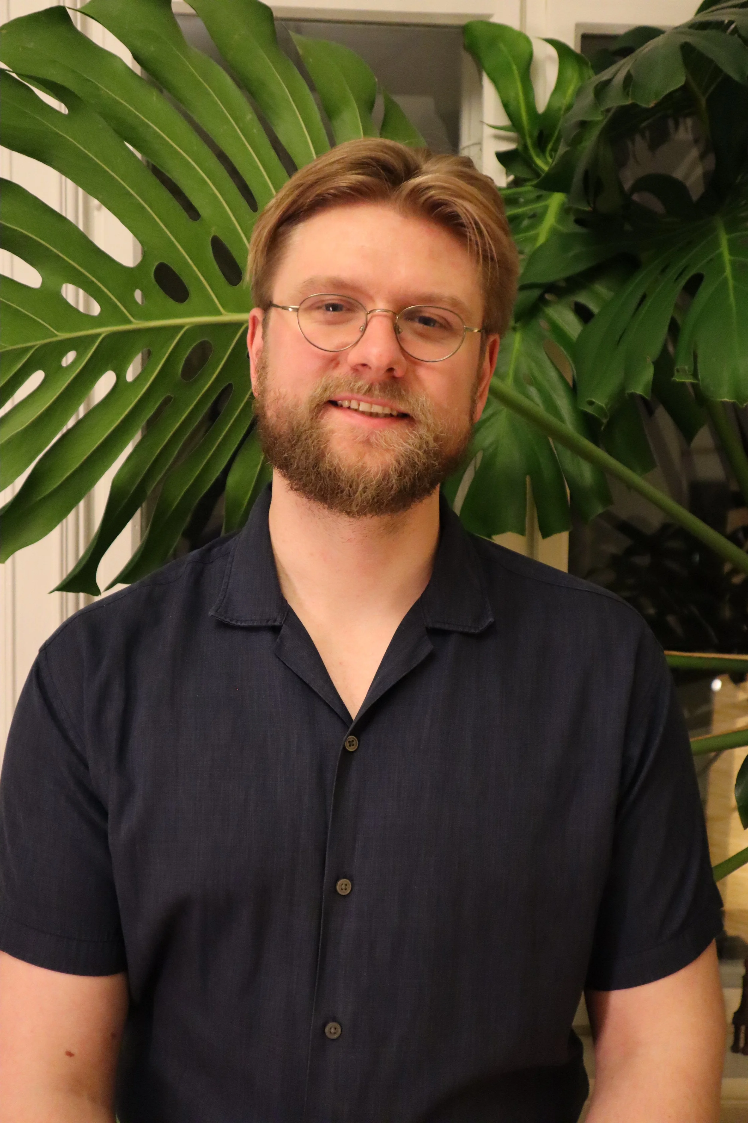 En mann med brunt hår, skjegg og briller som smiler foran grønne planter.