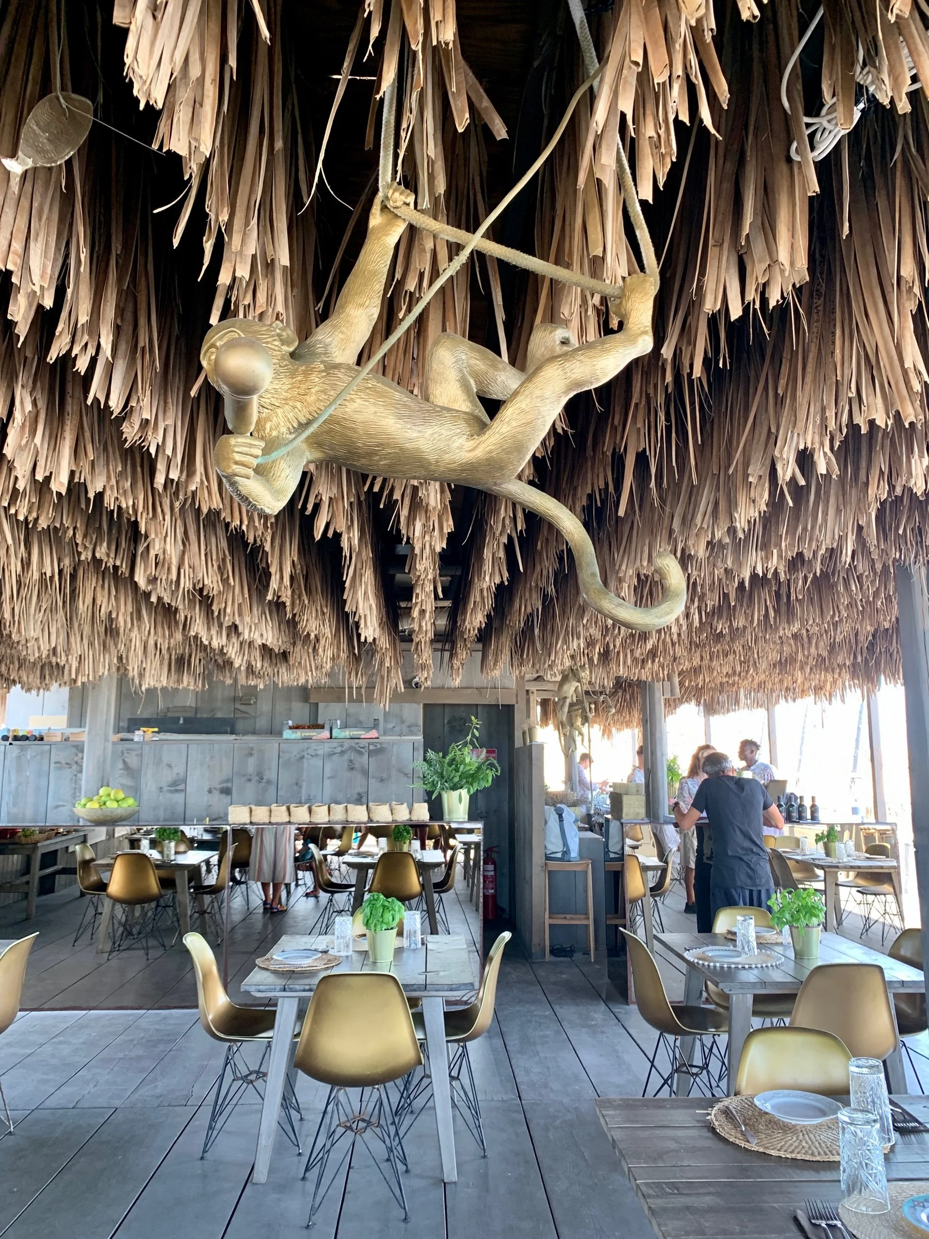 A restaurant interior with rustic decor featuring a thatched roof and a monkey sculpture hanging from the ceiling. The space is filled with tables and chairs, with some tables set with plates, glasses, and small plants. There are people standing and 