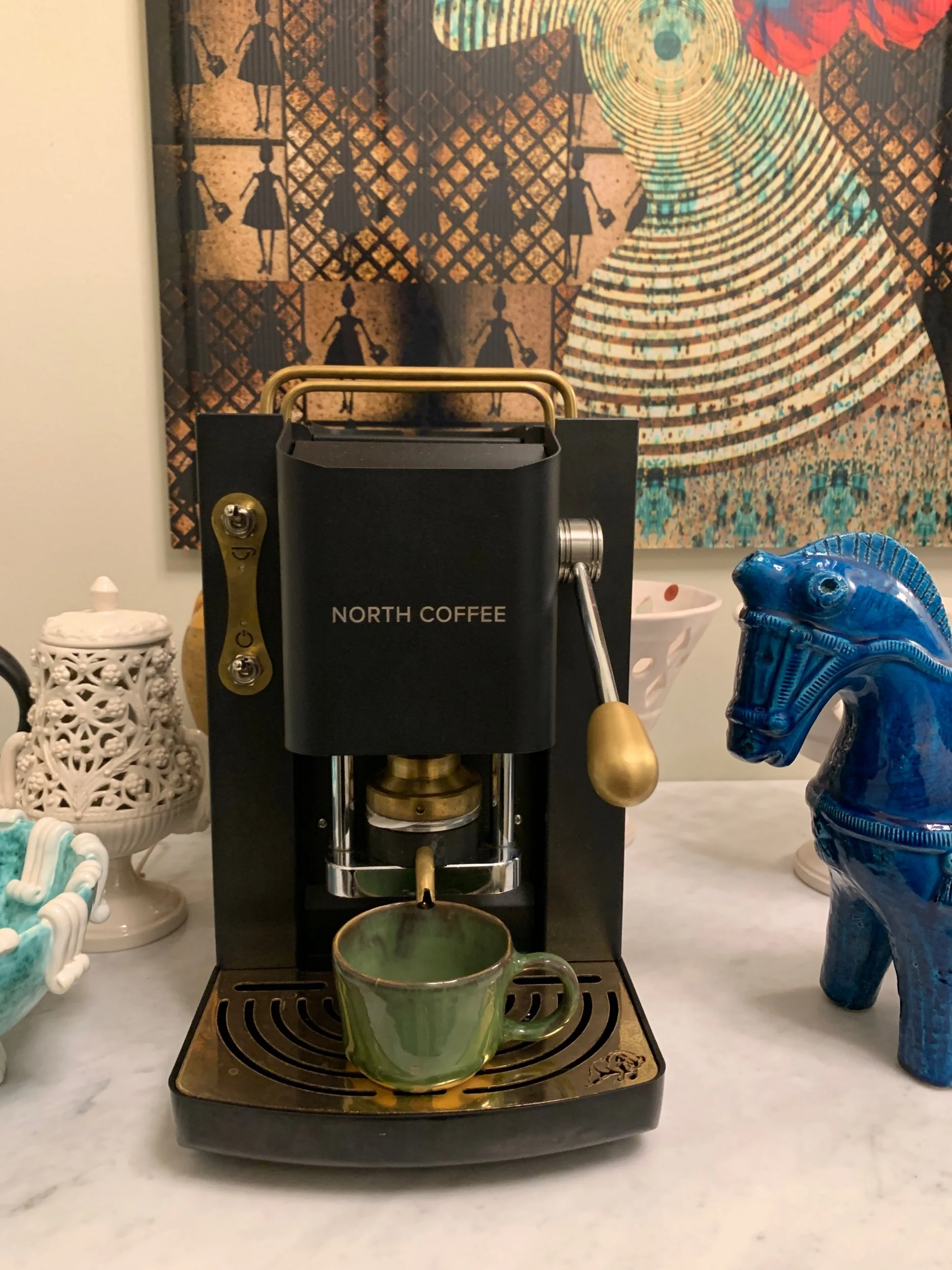 A coffee machine brewing into a green ceramic mug placed on its drip tray, with decorative items and artwork in the background.