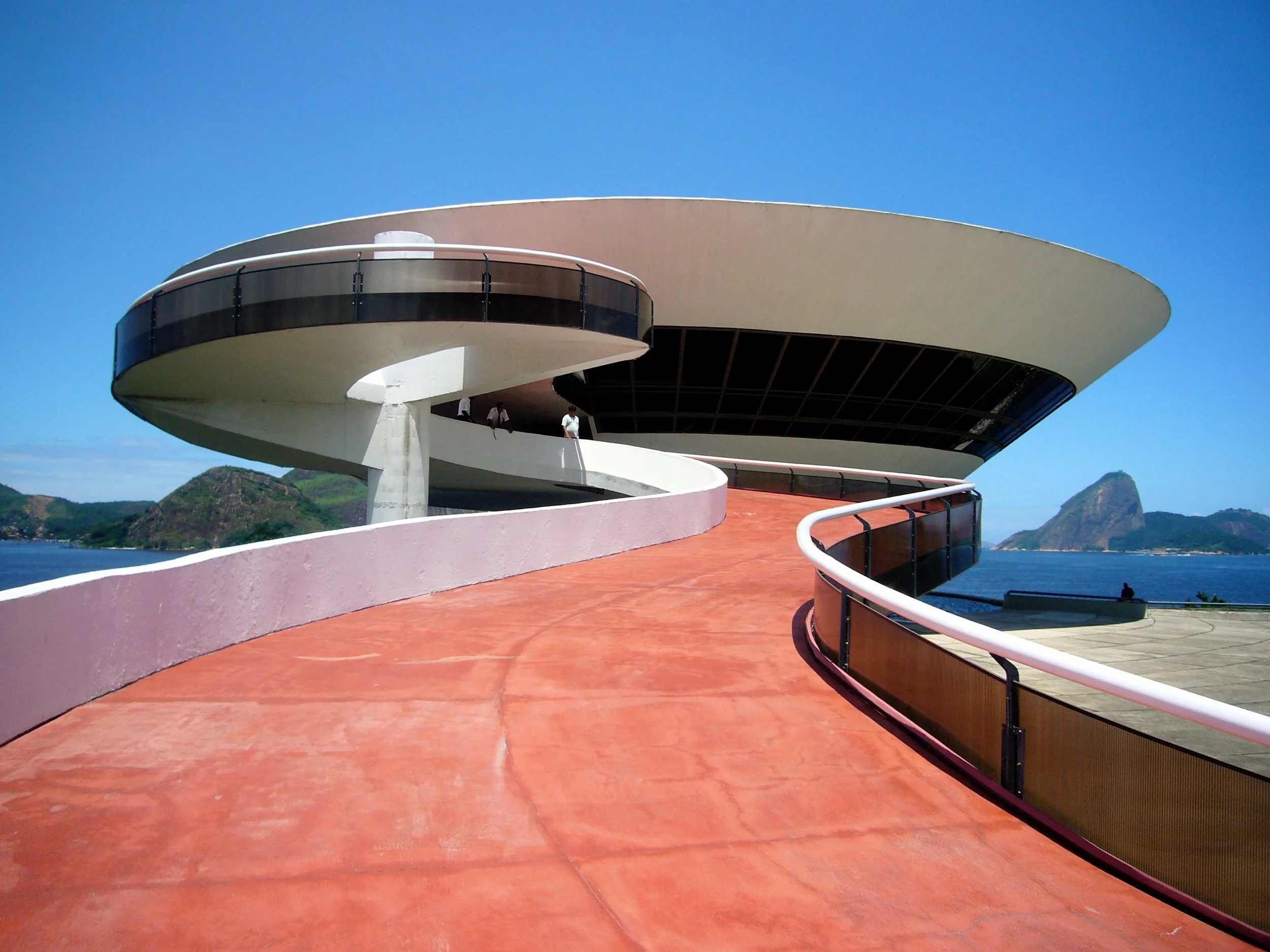 A modern, curved building with a large white roof, featuring a balcony with black glass railing, situated next to water and surrounded by green hills under a clear blue sky.