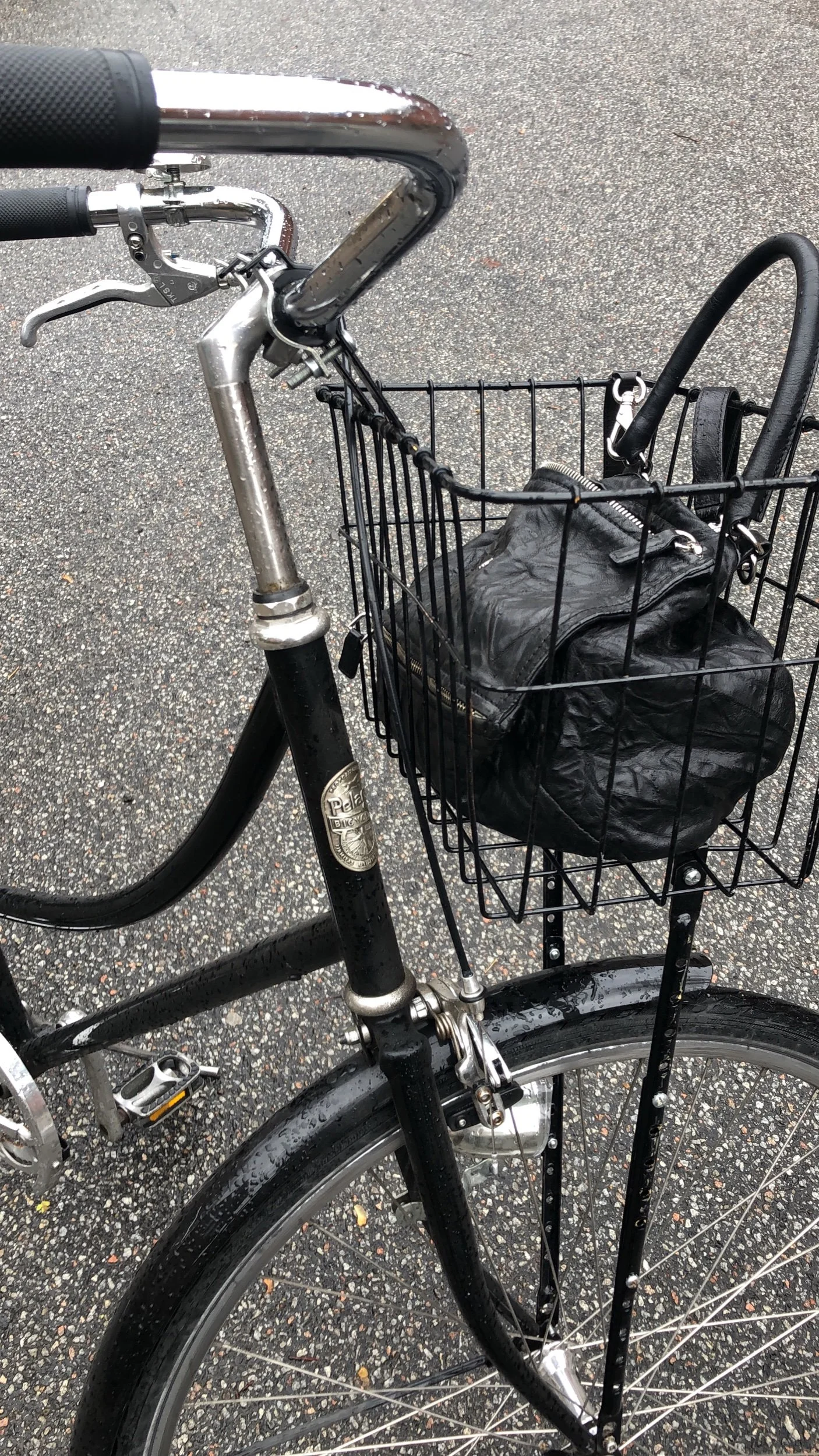 Close-up of a black bicycle with a front basket holding a black leather bag, on a wet asphalt road.