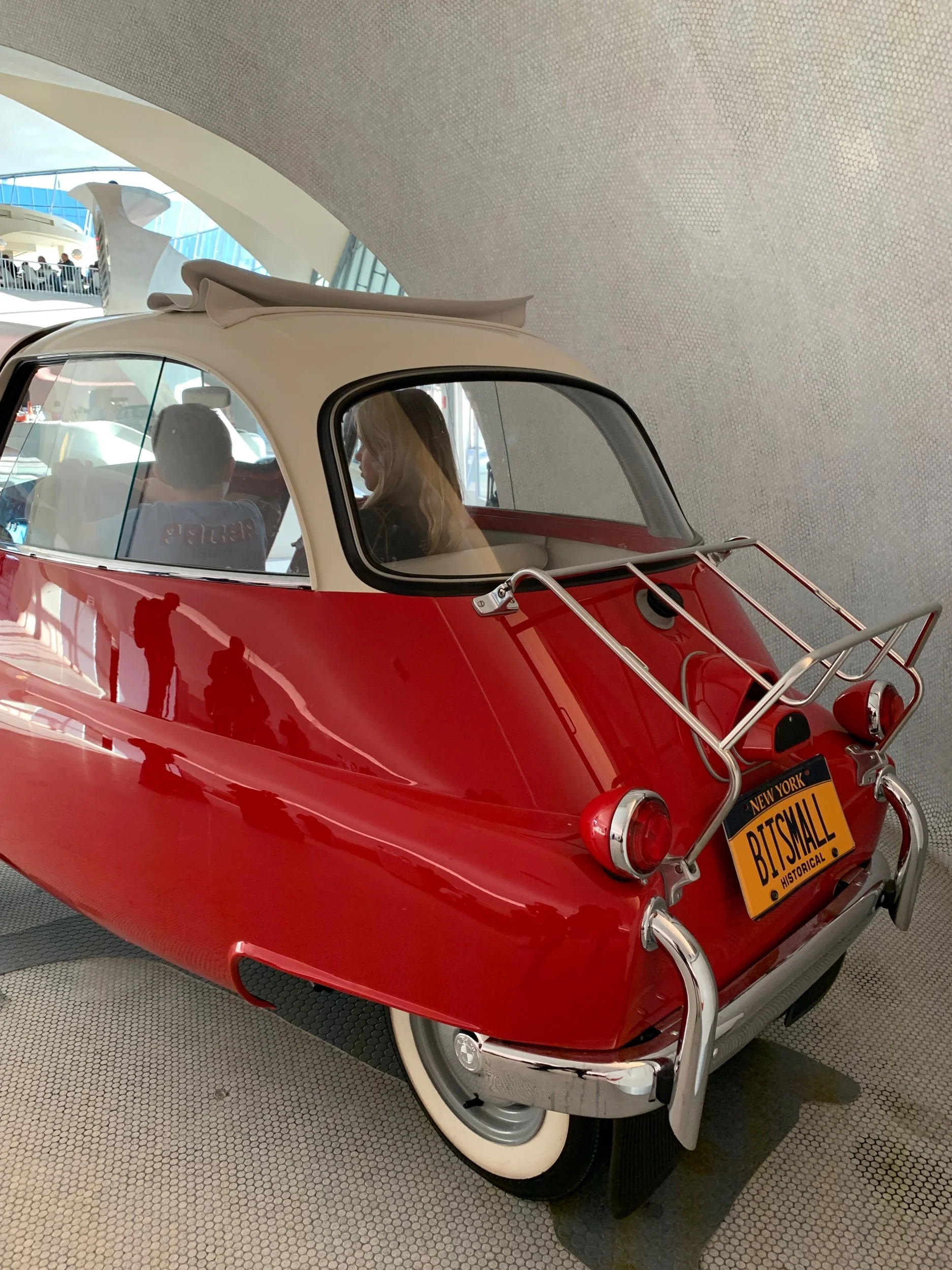 A vintage red and white bicycle with a 'BITSMAAL' license plate displayed in an indoor exhibit. The bike features a red body, white tires, and a beige canopy.