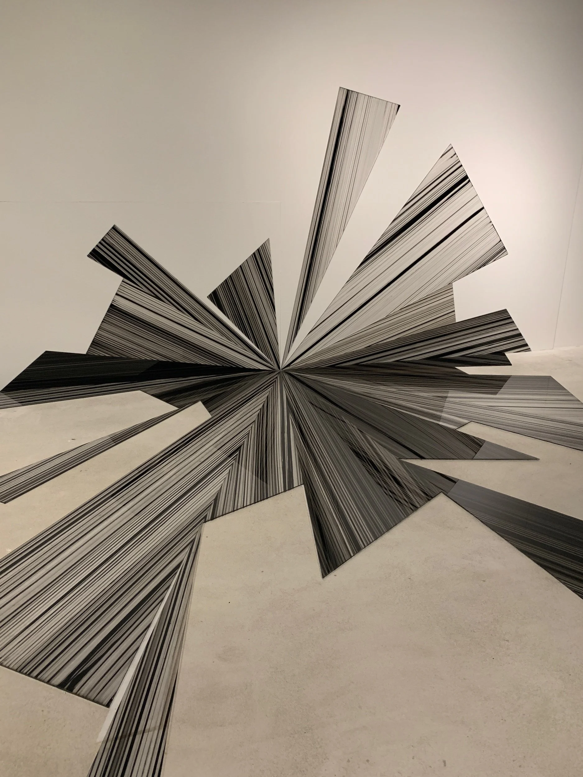 Art installation composed of numerous black and white striped paper or cardboard panels arranged in a fan shape on a white gallery wall.