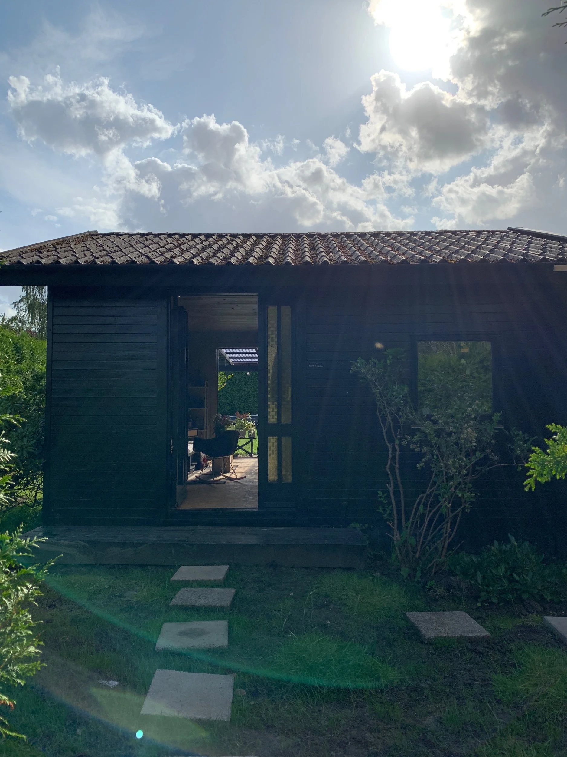 A small black wooden cabin with an open door, set in a green garden with stepping stones leading to the entrance, under a bright sunny sky with scattered clouds.