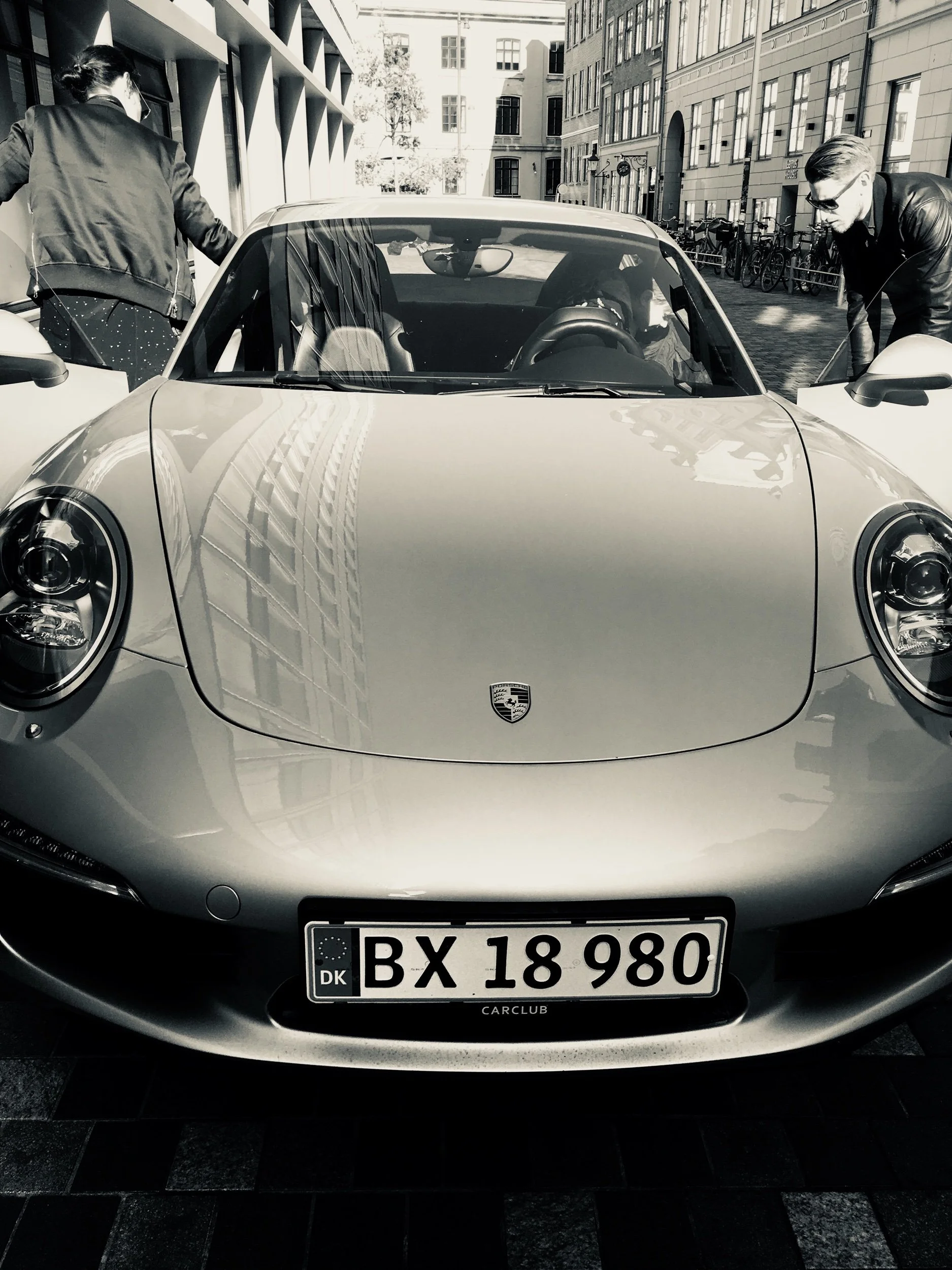 Front view of a silver Porsche sports car with Danish license plates, parked on a city street. Two people are examining the car, one on each side. The street has modern buildings and bicycles in the background.