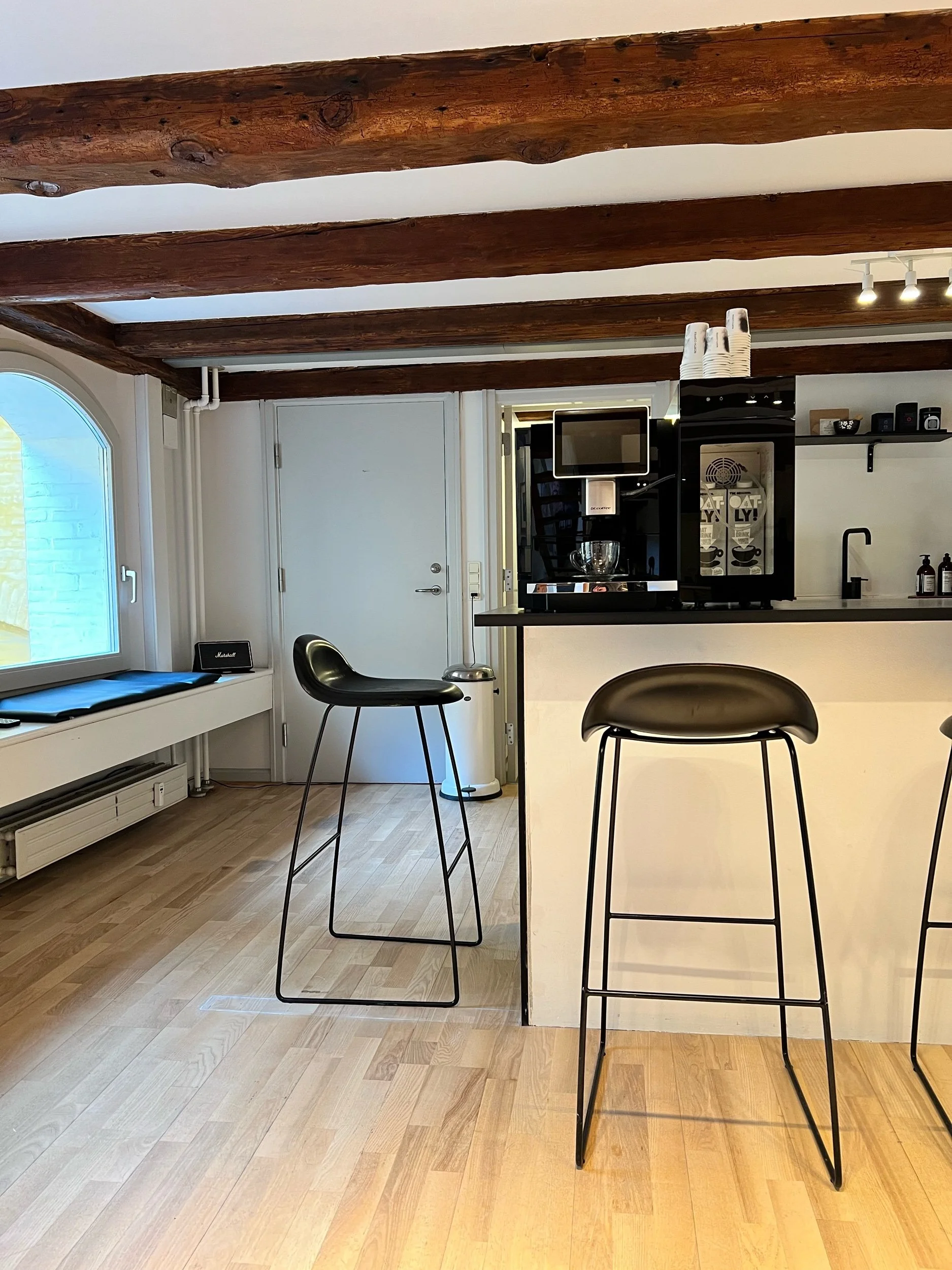 Interior of a modern kitchen with wooden beams on the ceiling, a window on the left, and black bar stools in the foreground. There's a coffee machine and a small shelf with various items on the counter.