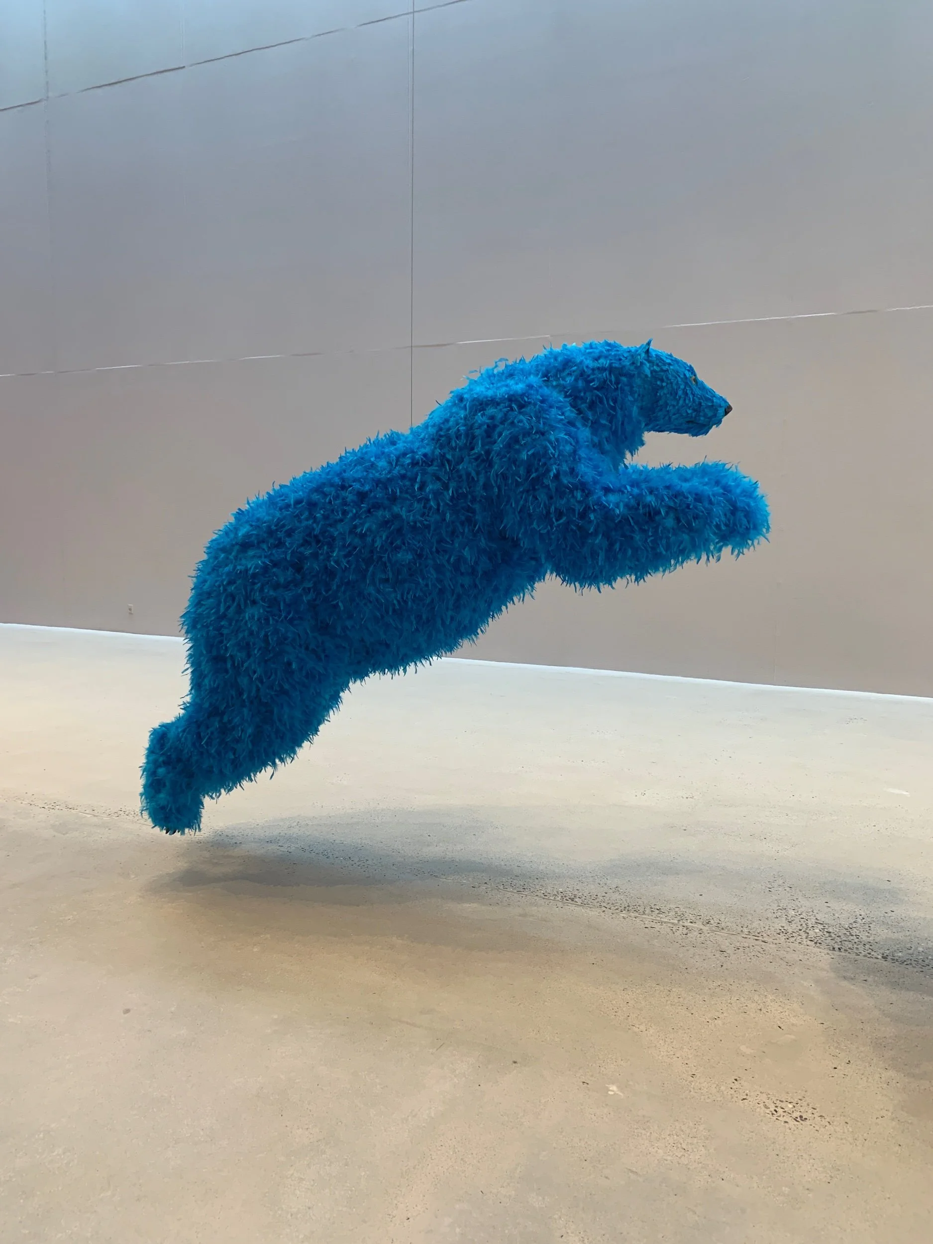 Large blue furry animal sculpture suspended in mid-air in a gallery with plain gray walls.
