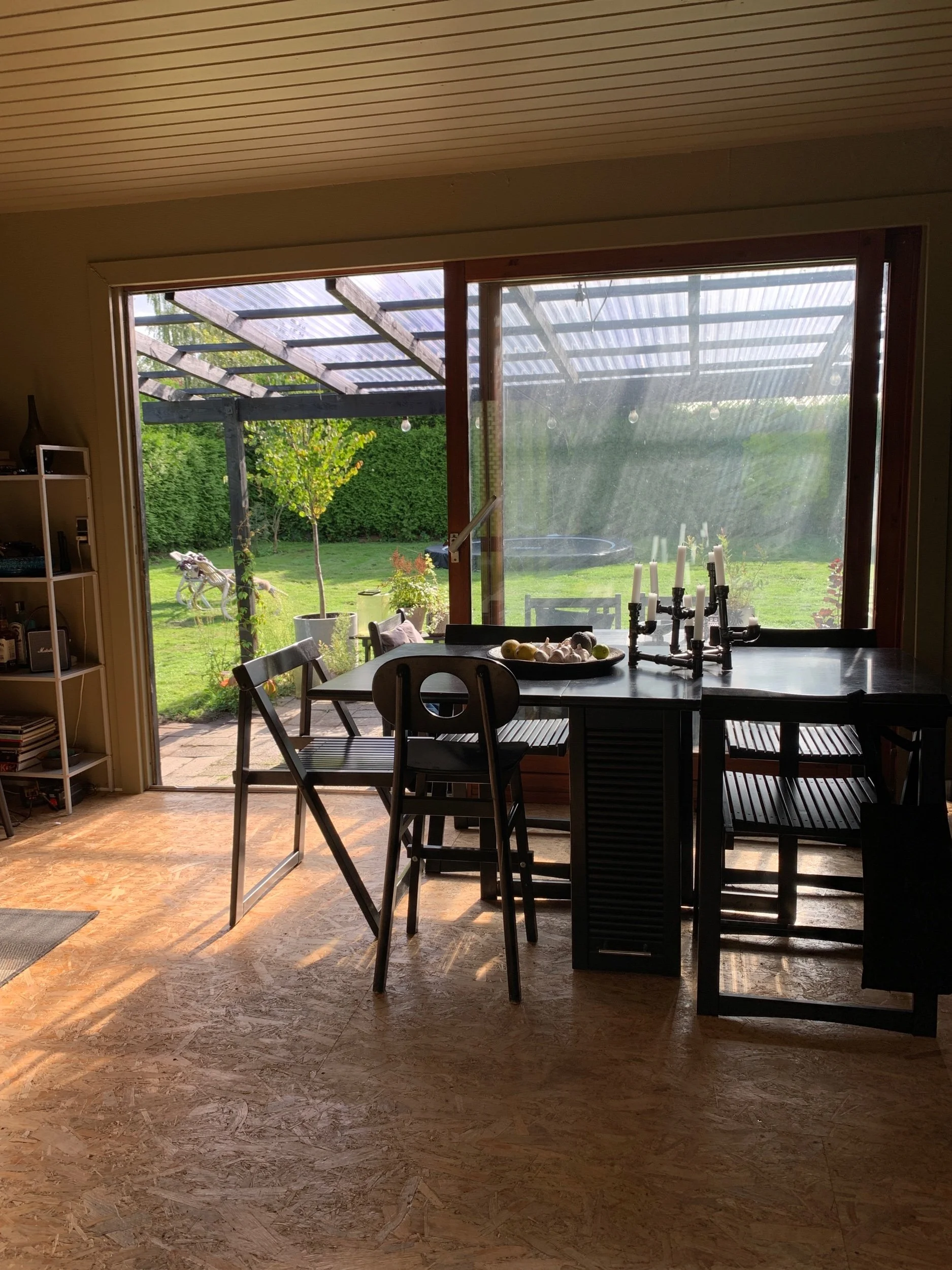 Interior view of a dining area with a large window showing a backyard garden with trees and a lawn, natural sunlight coming in, a black table with chairs, a candelabra, and a round tray of apples and garlics on the table.